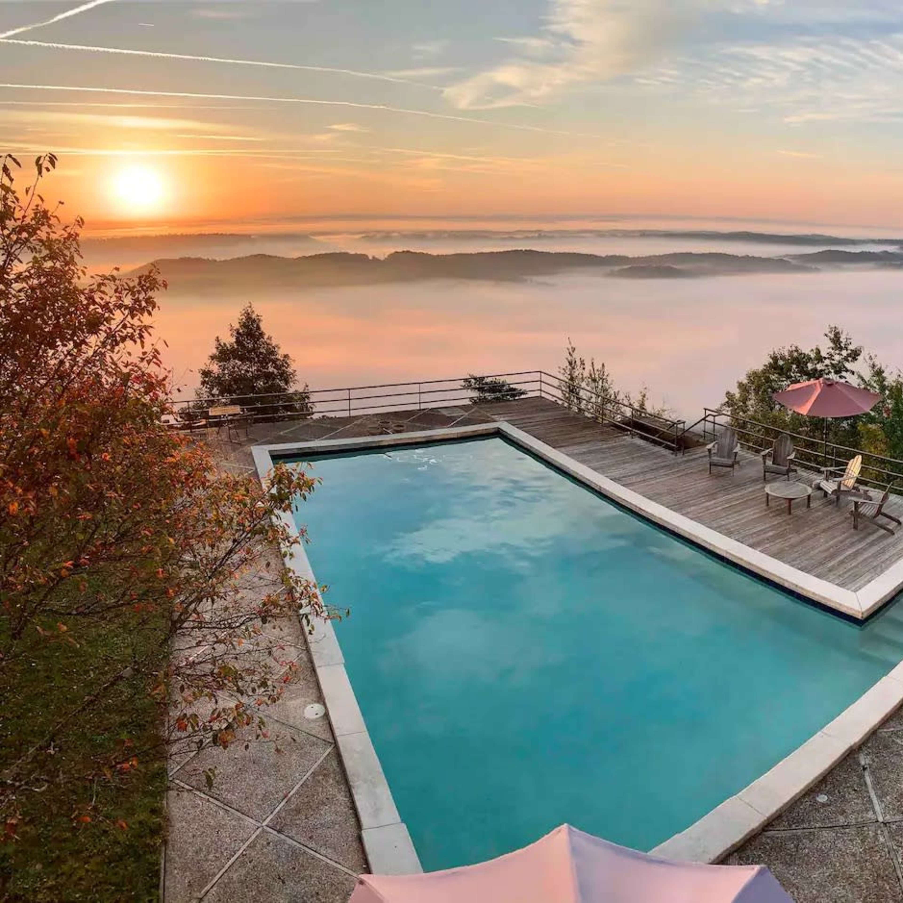 A rectangular swimming pool is surrounded by a wooden deck, overlooking a landscape of misty hills at sunrise.