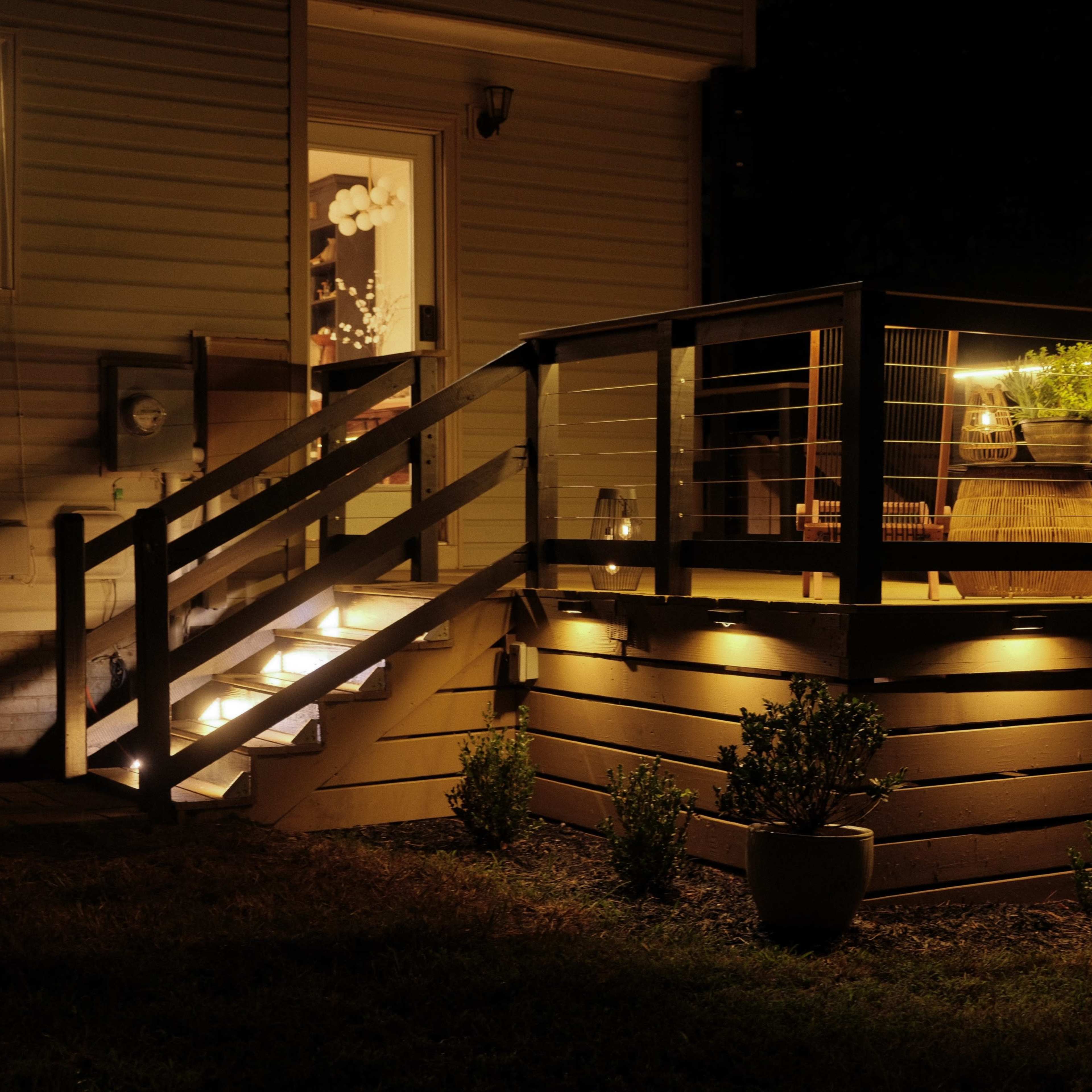 A wooden deck with railing and stairs is illuminated by lights at night, showcasing potted plants and a small table.