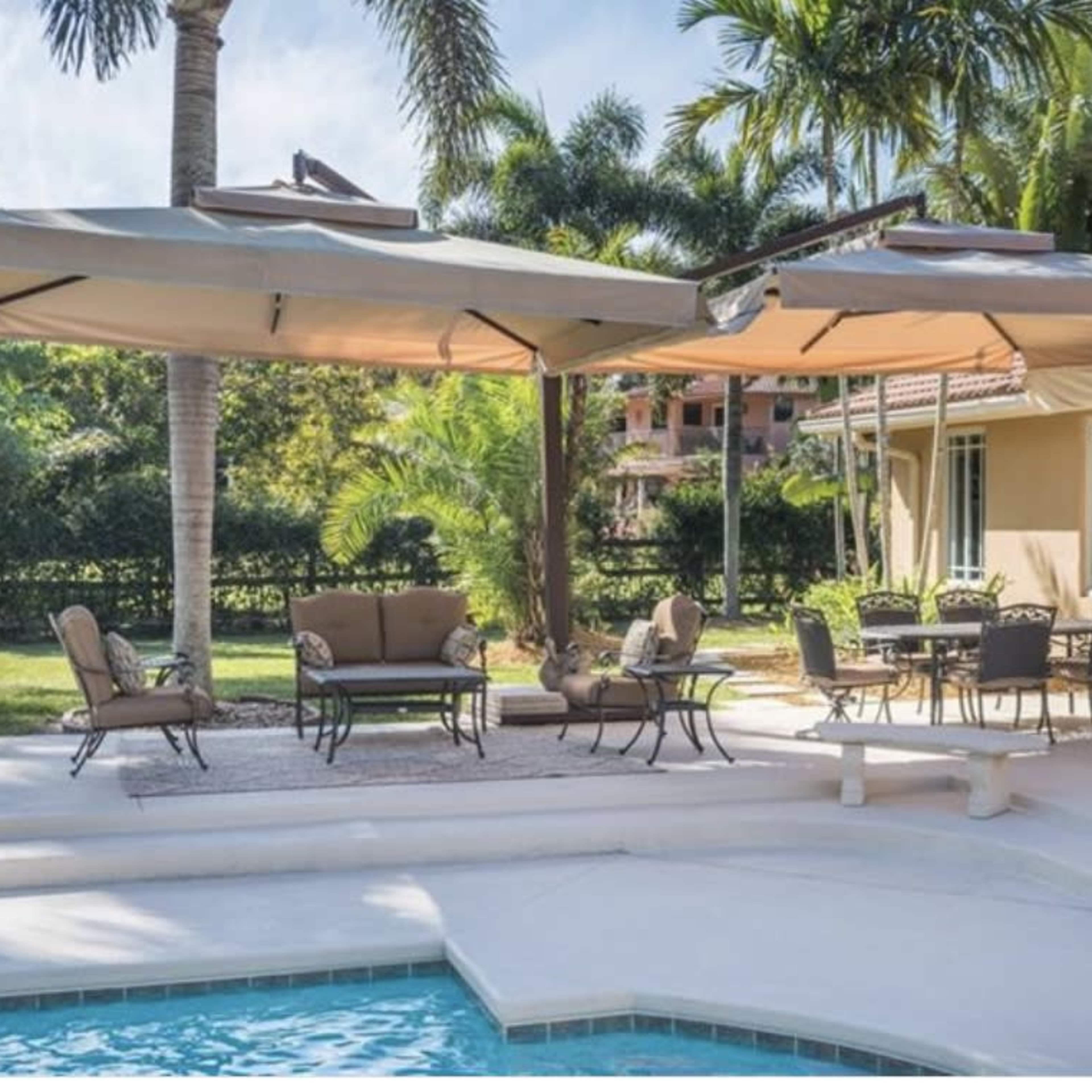 A patio area with a pool features a seating arrangement under large umbrellas and a dining table surrounded by palm trees.