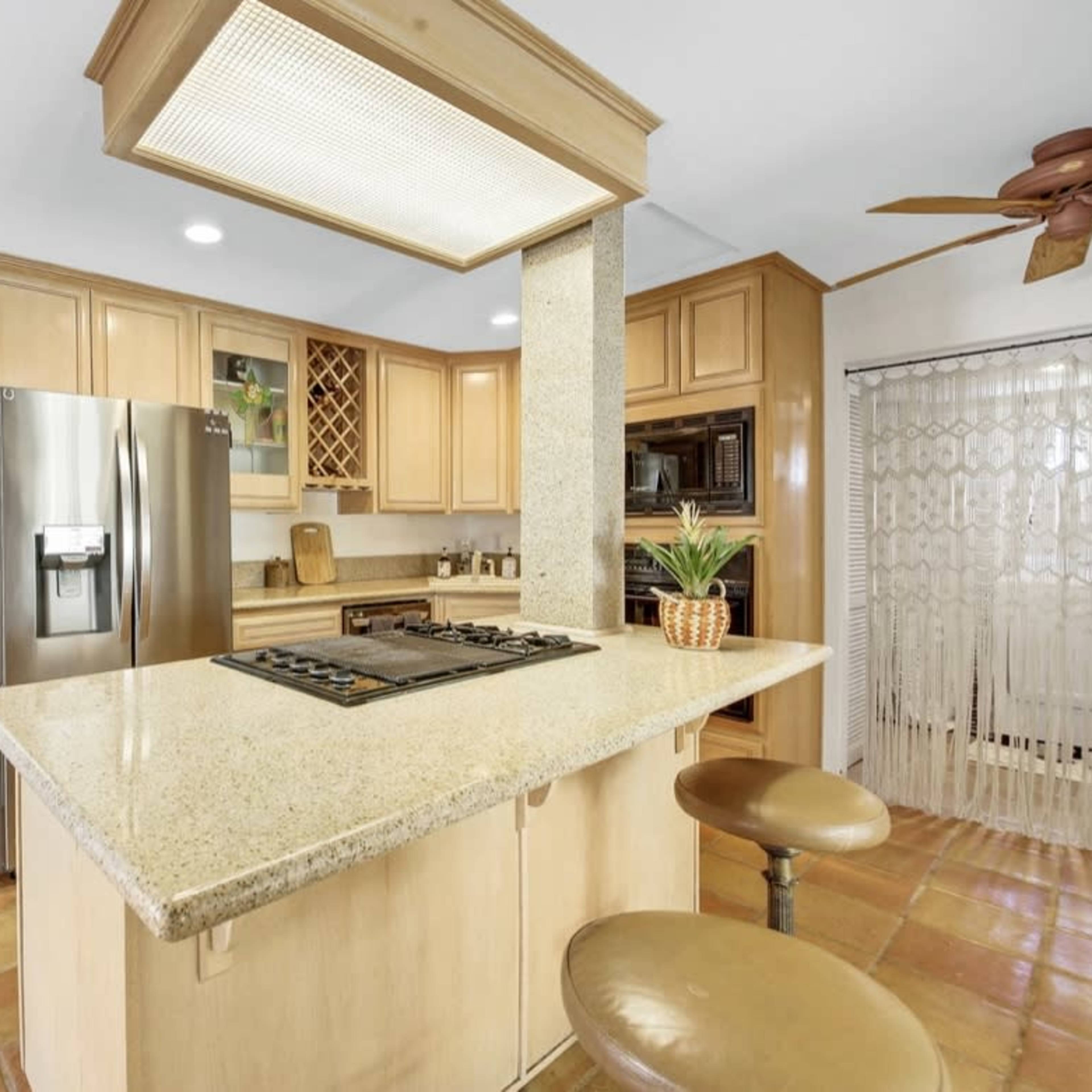 A kitchen with wooden cabinets, a central island with a gas cooktop, and modern appliances, featuring tile flooring and a doorway leading to another room.