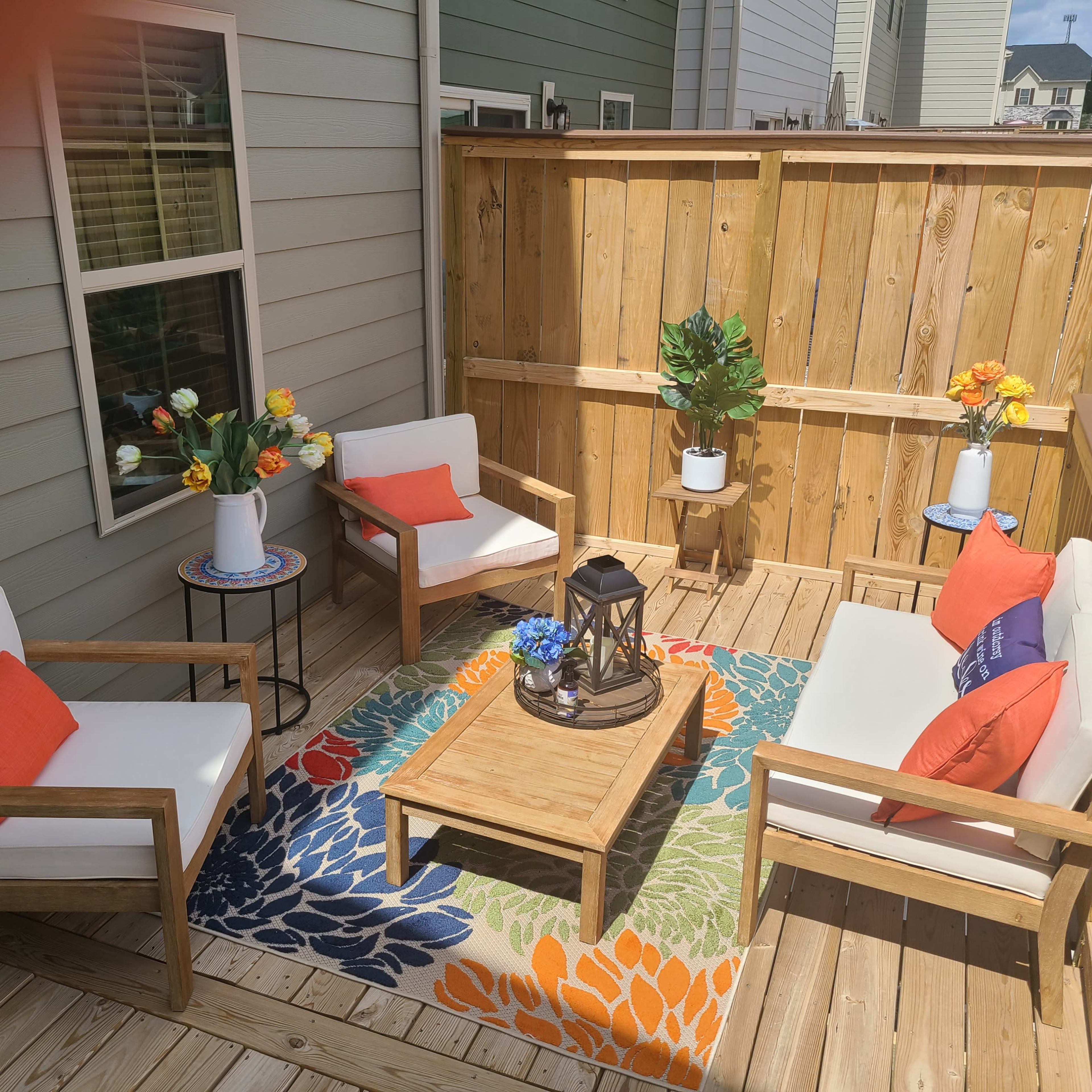 A small outdoor balcony with two armchairs, a coffee table, a colorful rug, and decorative plants surrounded by wooden fencing.