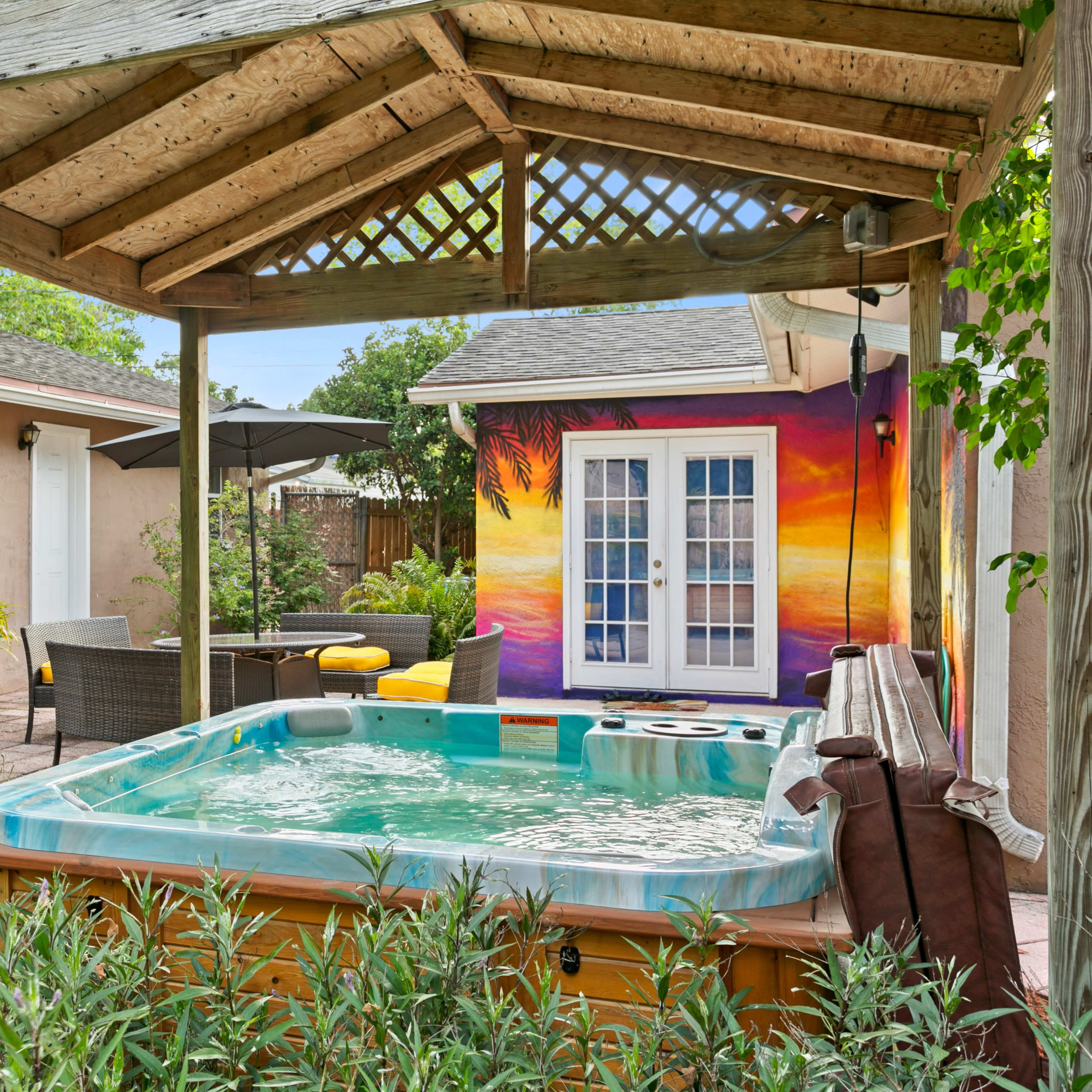 A hot tub sits under a wooden gazebo in a backyard with tropical plants and a colorful mural on the wall of a nearby building.