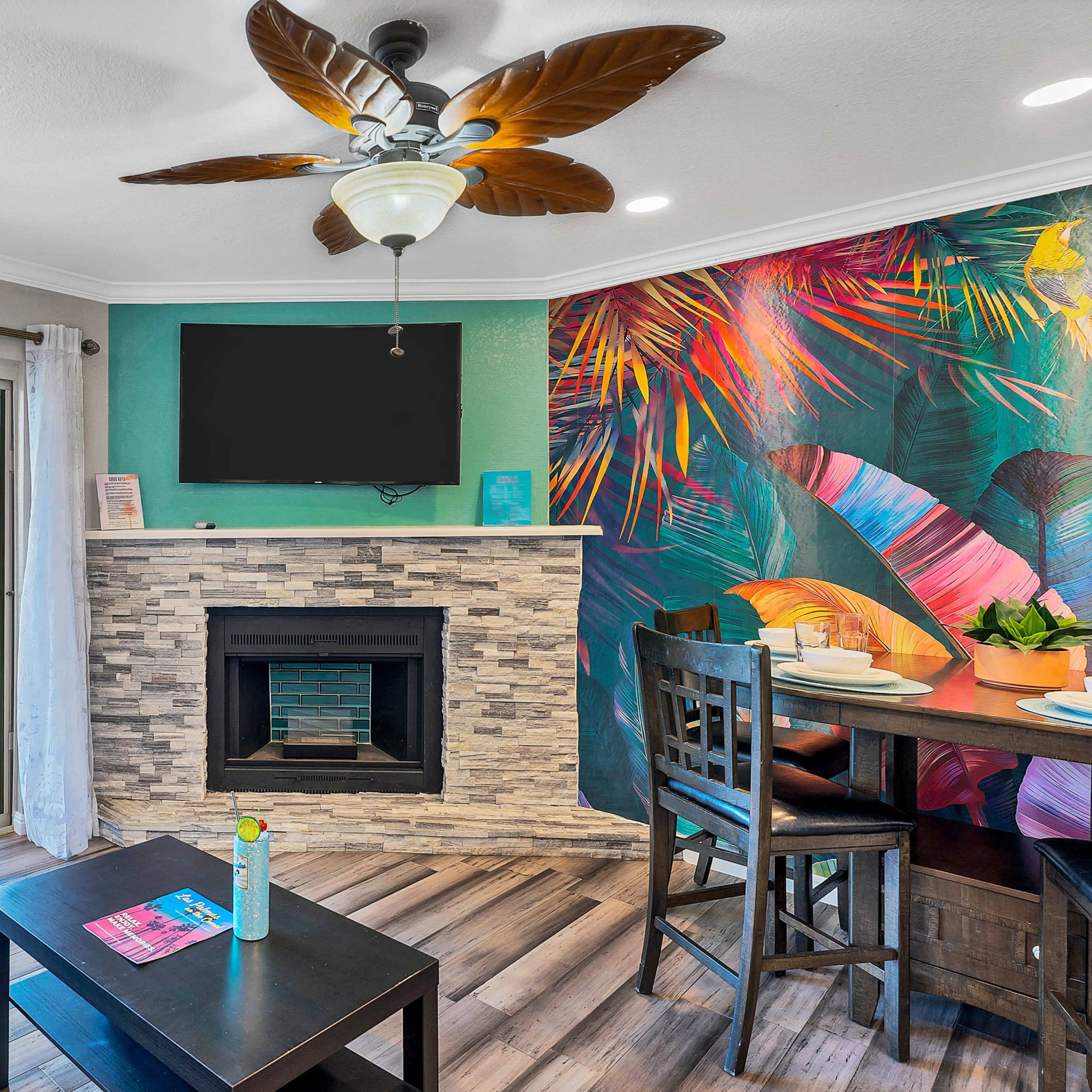 A living area featuring a dark leather sofa, a stone fireplace, a dining table, and a vibrant mural with tropical foliage, alongside a balcony door leading to an outdoor view.