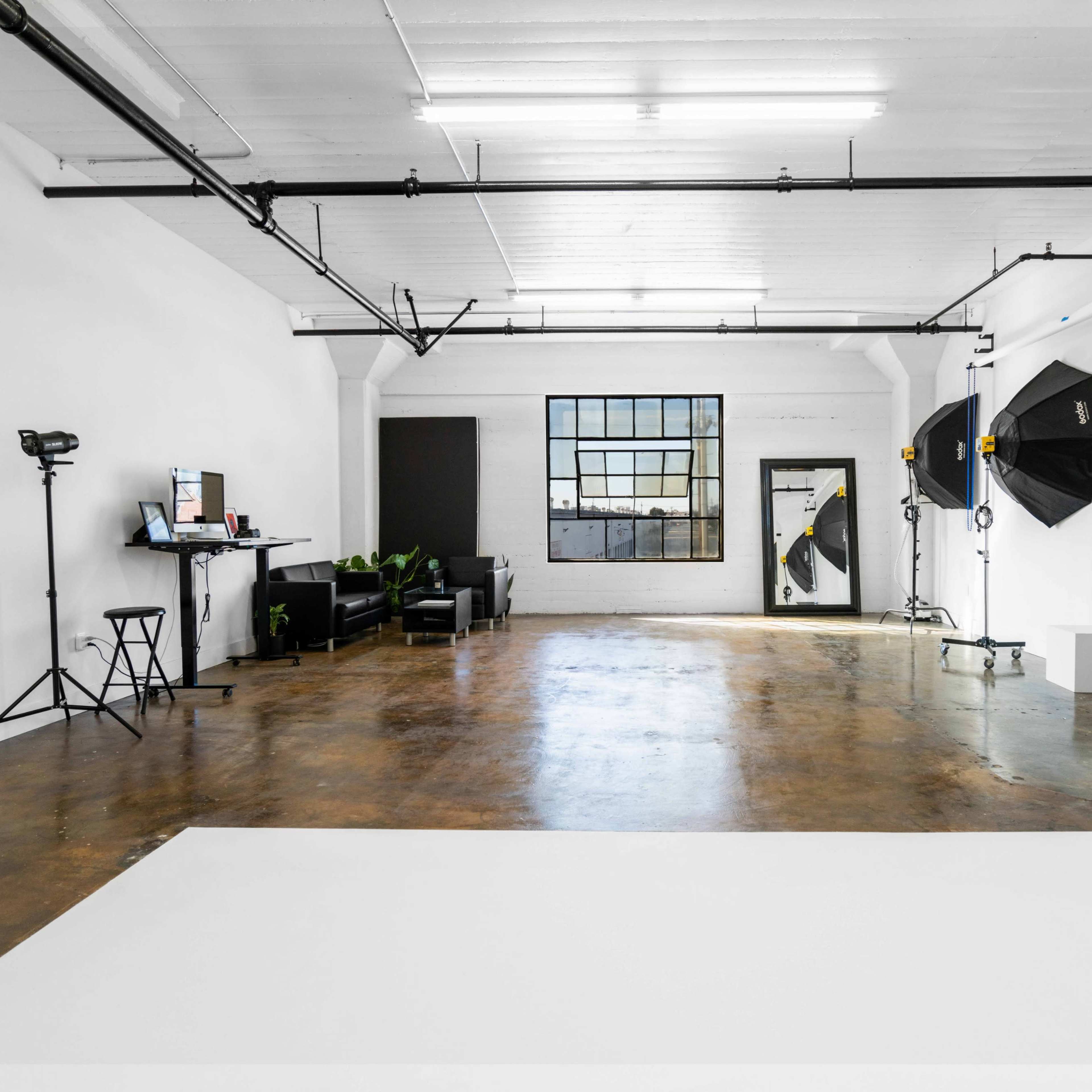 A spacious studio with a polished concrete floor, illuminated by softbox lights, a large window, and minimal furniture.