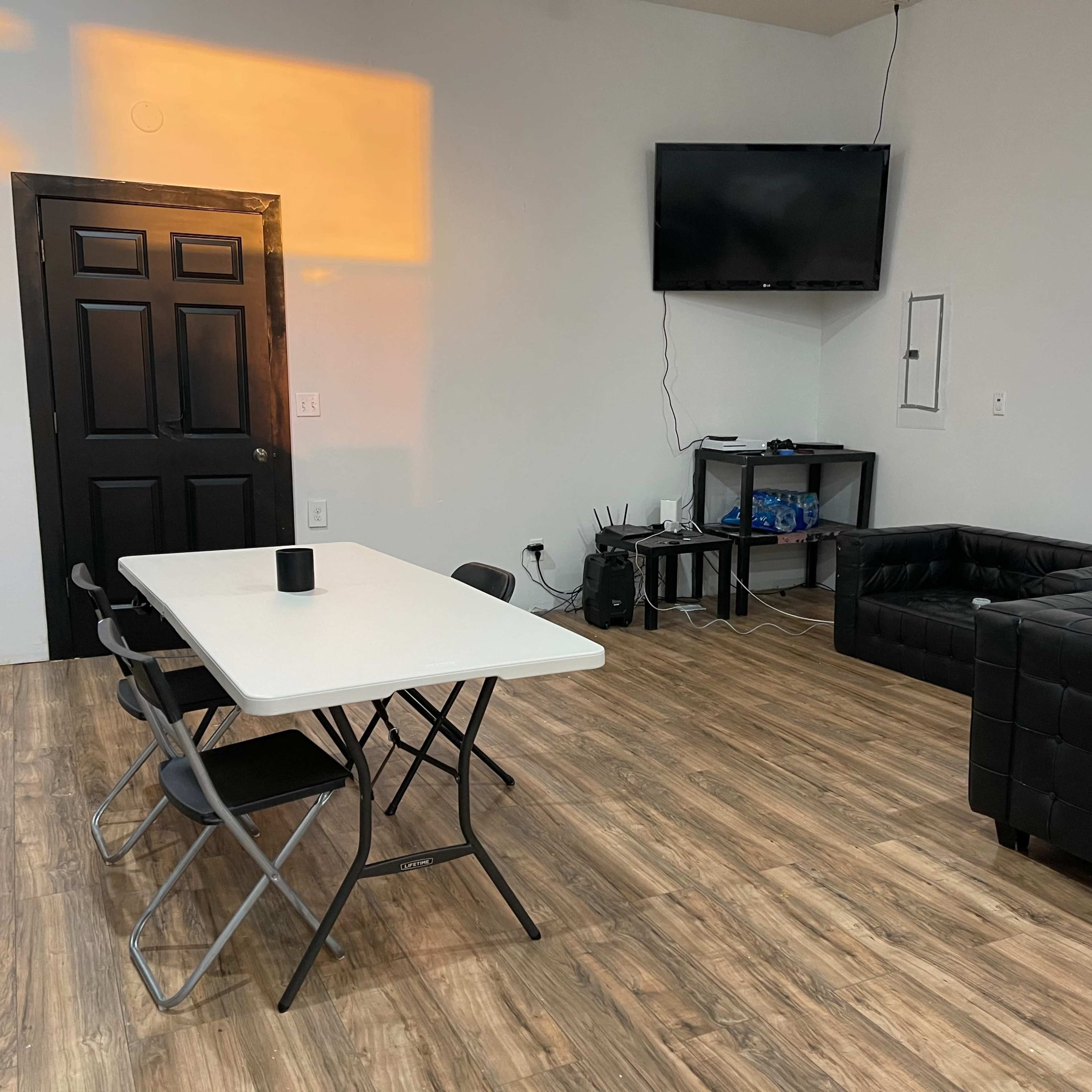 A simple room with a black table and chairs, a black leather couch, a television on the wall, and a small entertainment unit against the wall.