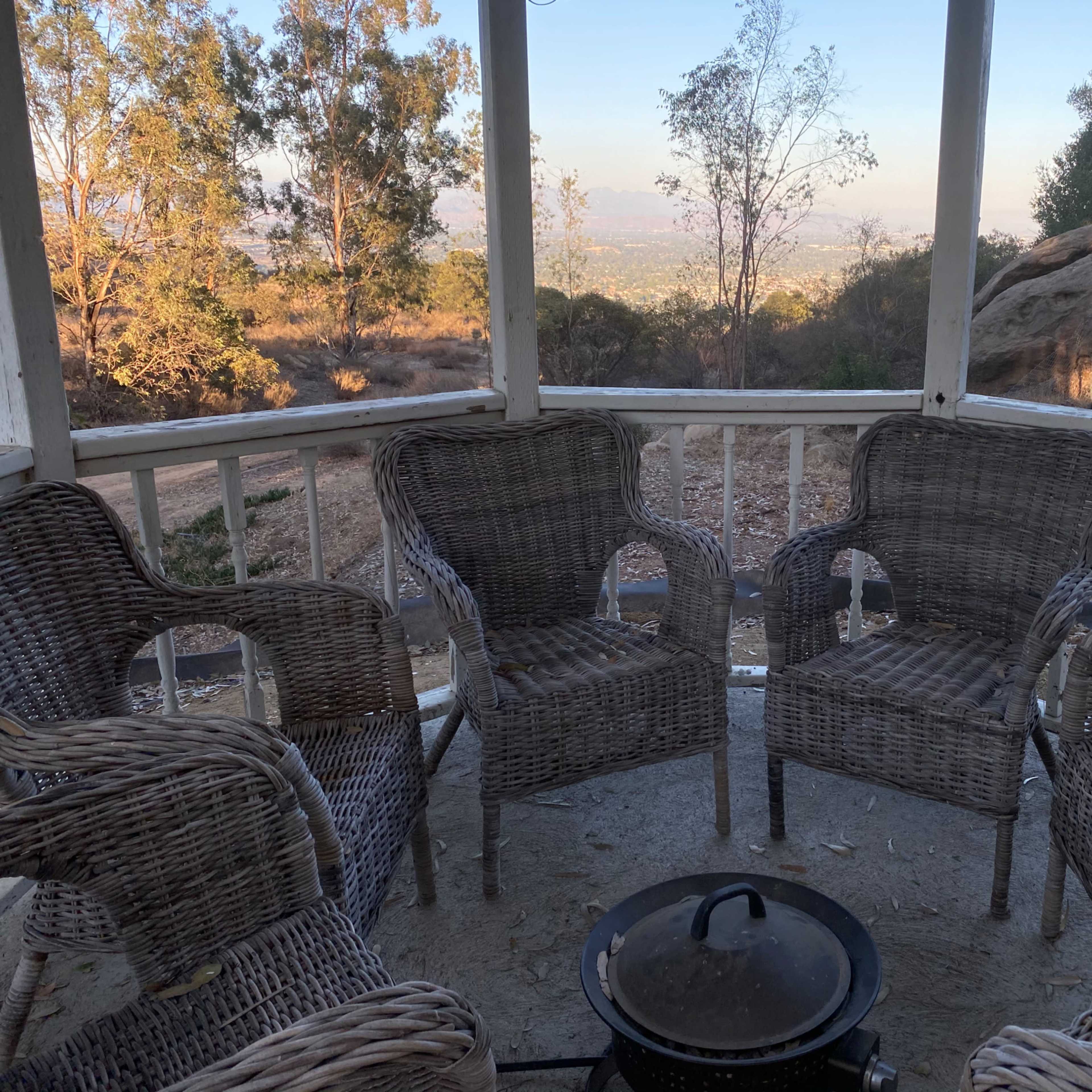 A circular patio with four wicker chairs surrounds a black pot in an open gazebo, overlooking a valley filled with trees and distant hills.