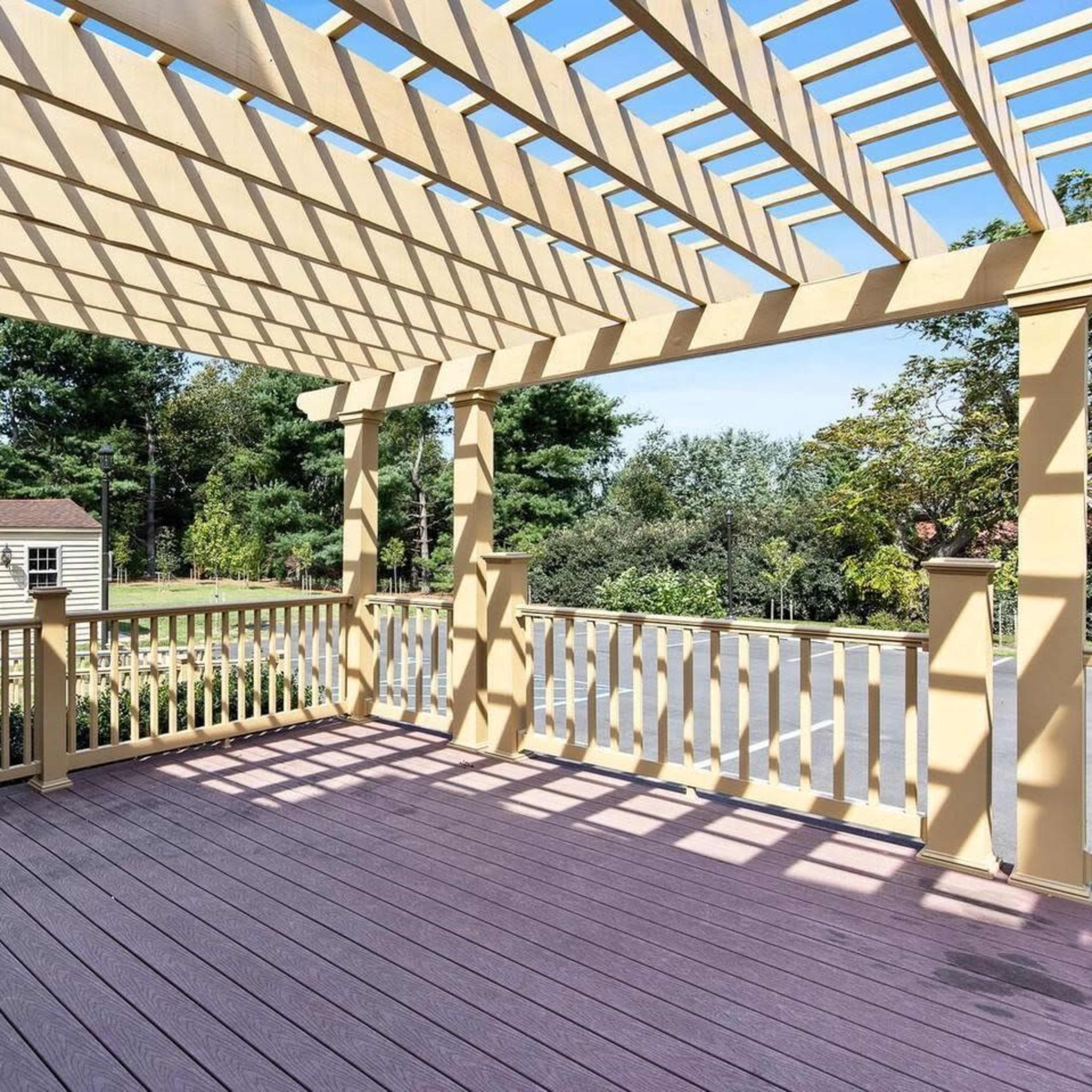 A wooden deck with a pergola structure above, surrounded by trees and a nearby shed in the background.