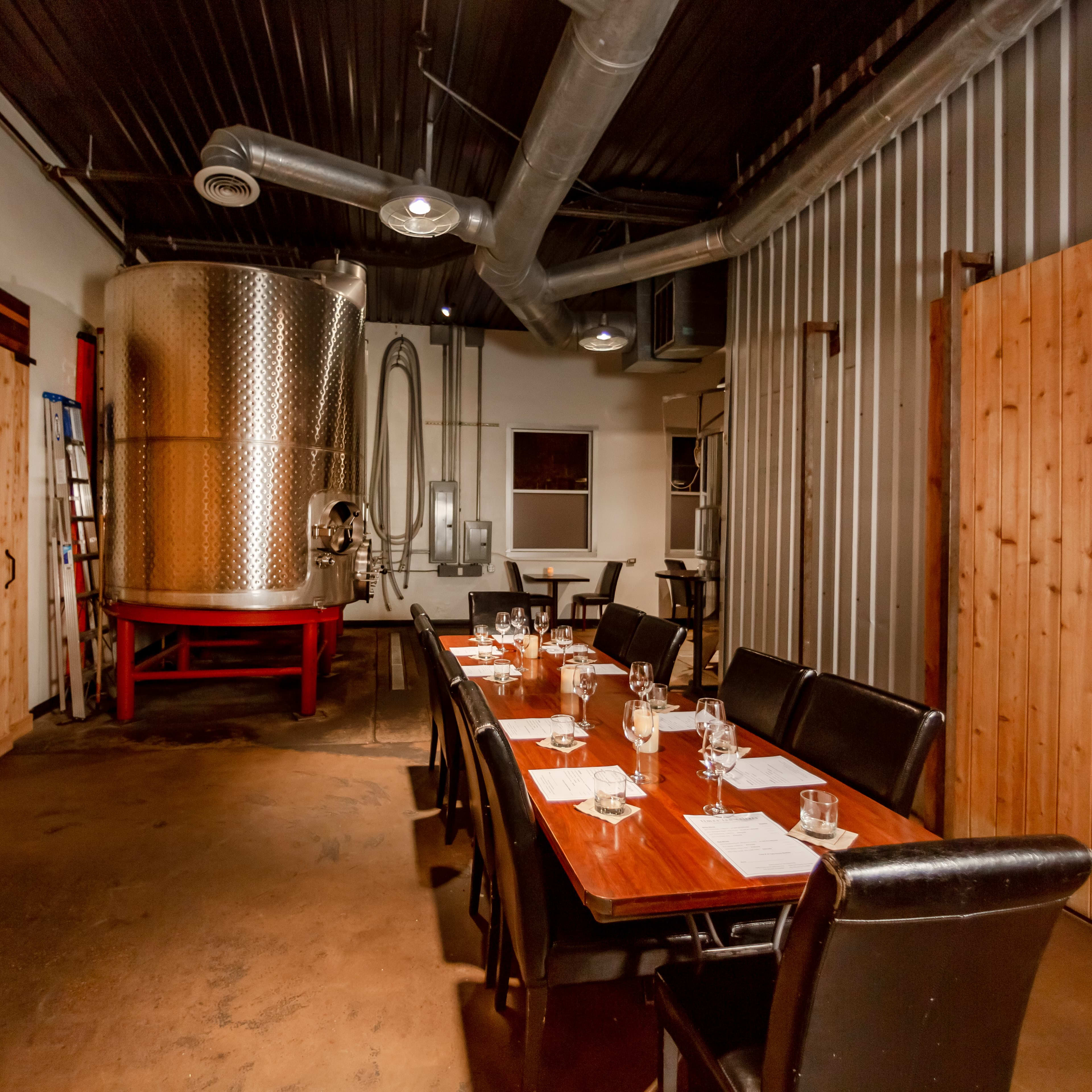 A dining area with a long wooden table set for a meal, adjacent to a large metal fermentation tank and wooden walls.