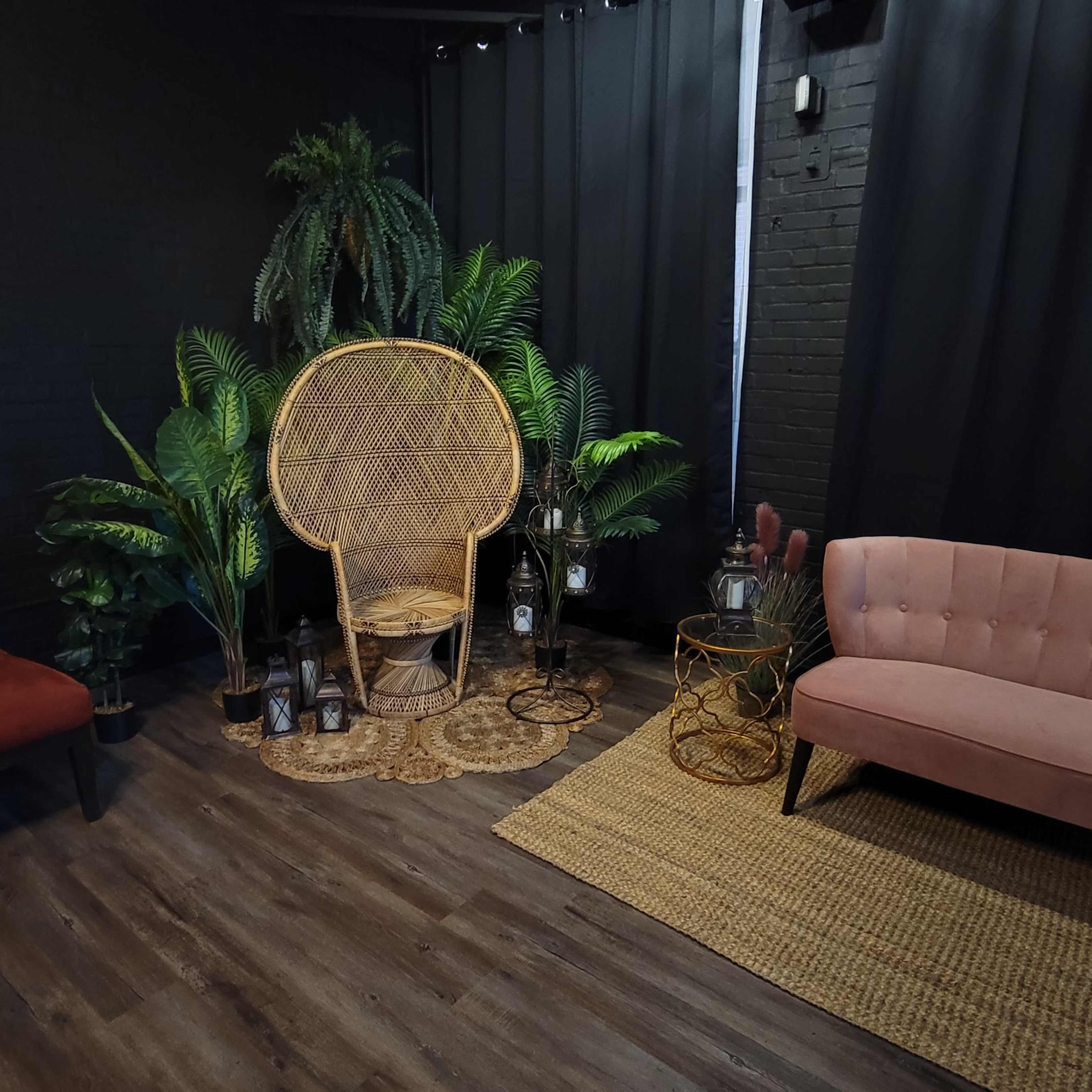 A cozy indoor space with a rattan chair surrounded by potted plants, a pink velvet couch, and a red armchair on a wooden floor.