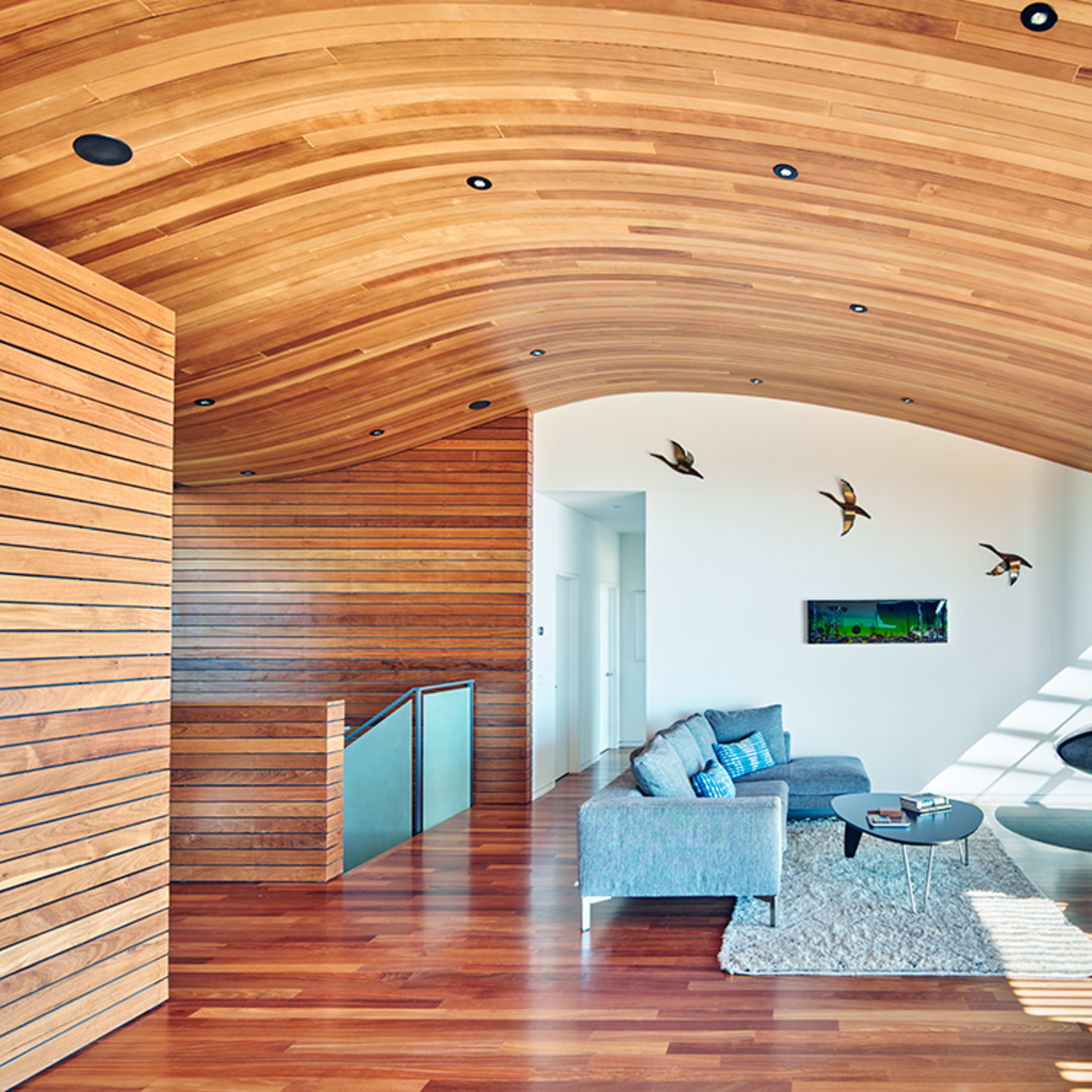 A modern living room with a curved wooden ceiling, hardwood floors, and a sleek fireplace, featuring a sectional sofa, a coffee table, and a view of the outdoors through large glass doors.