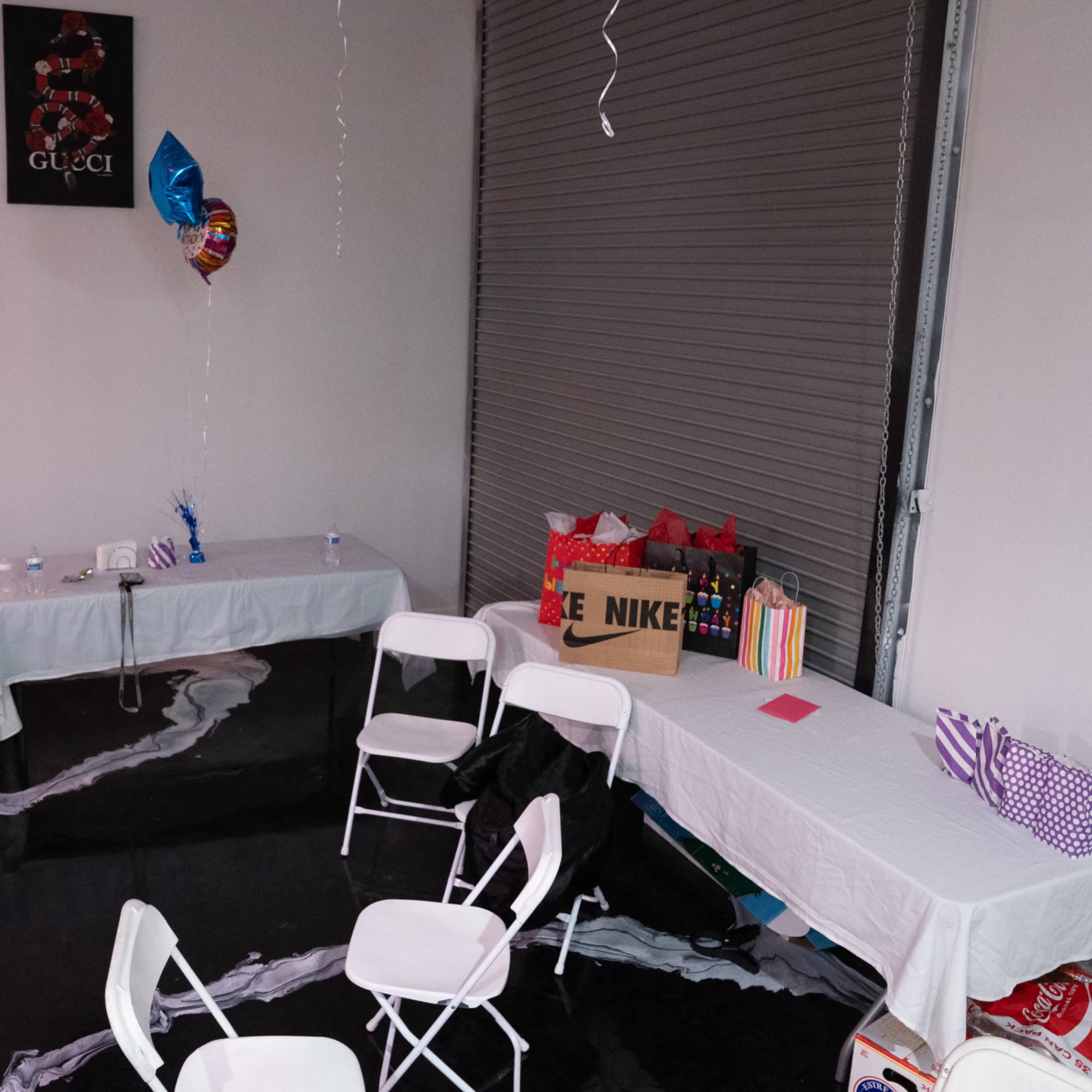 A party setup with white tables and chairs, decorated with balloons, and gifts stored on a shelf against a wall.