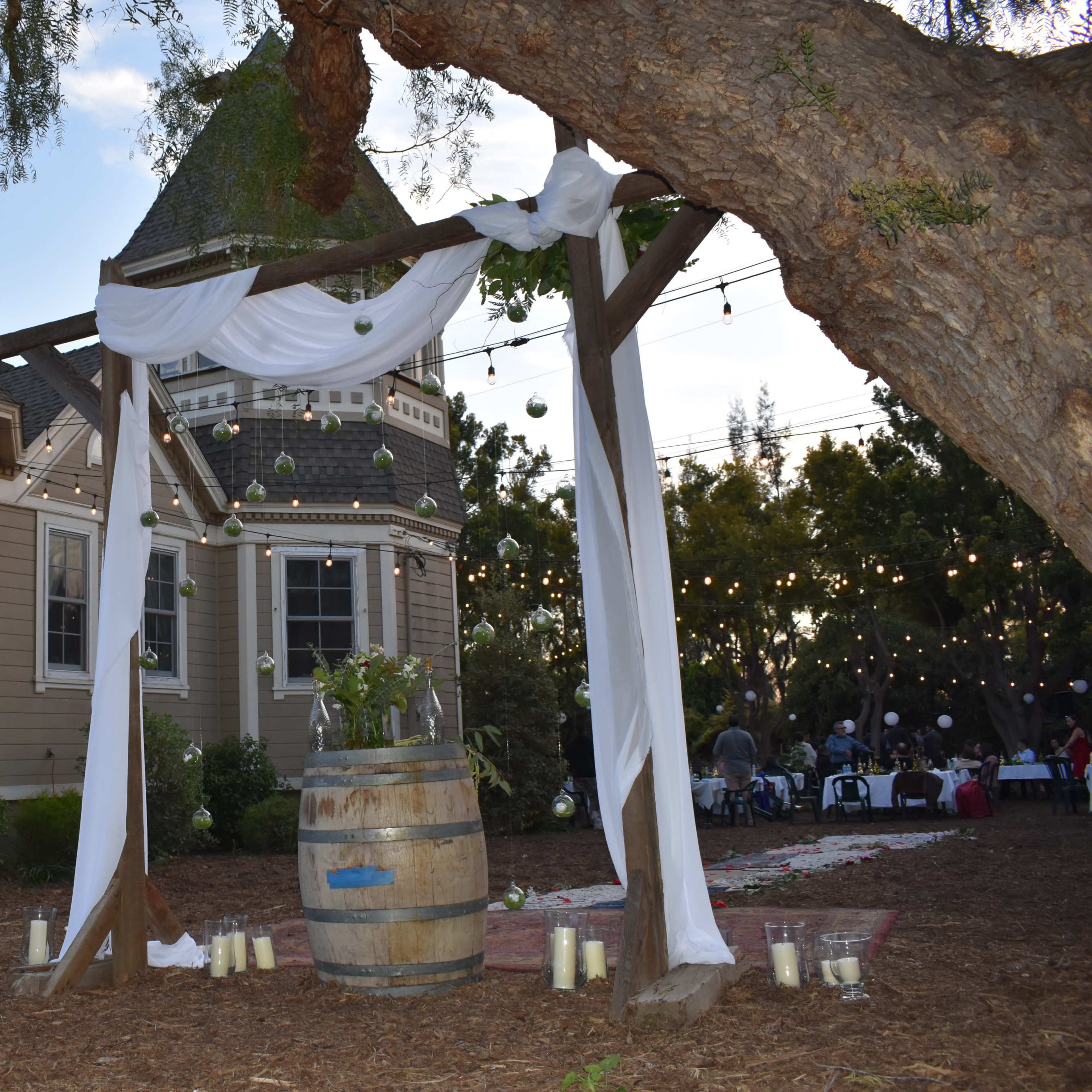 An outdoor wedding setup features a wooden arch draped with fabric, standing beside a large barrel, with string lights illuminating the scene behind a quaint house.