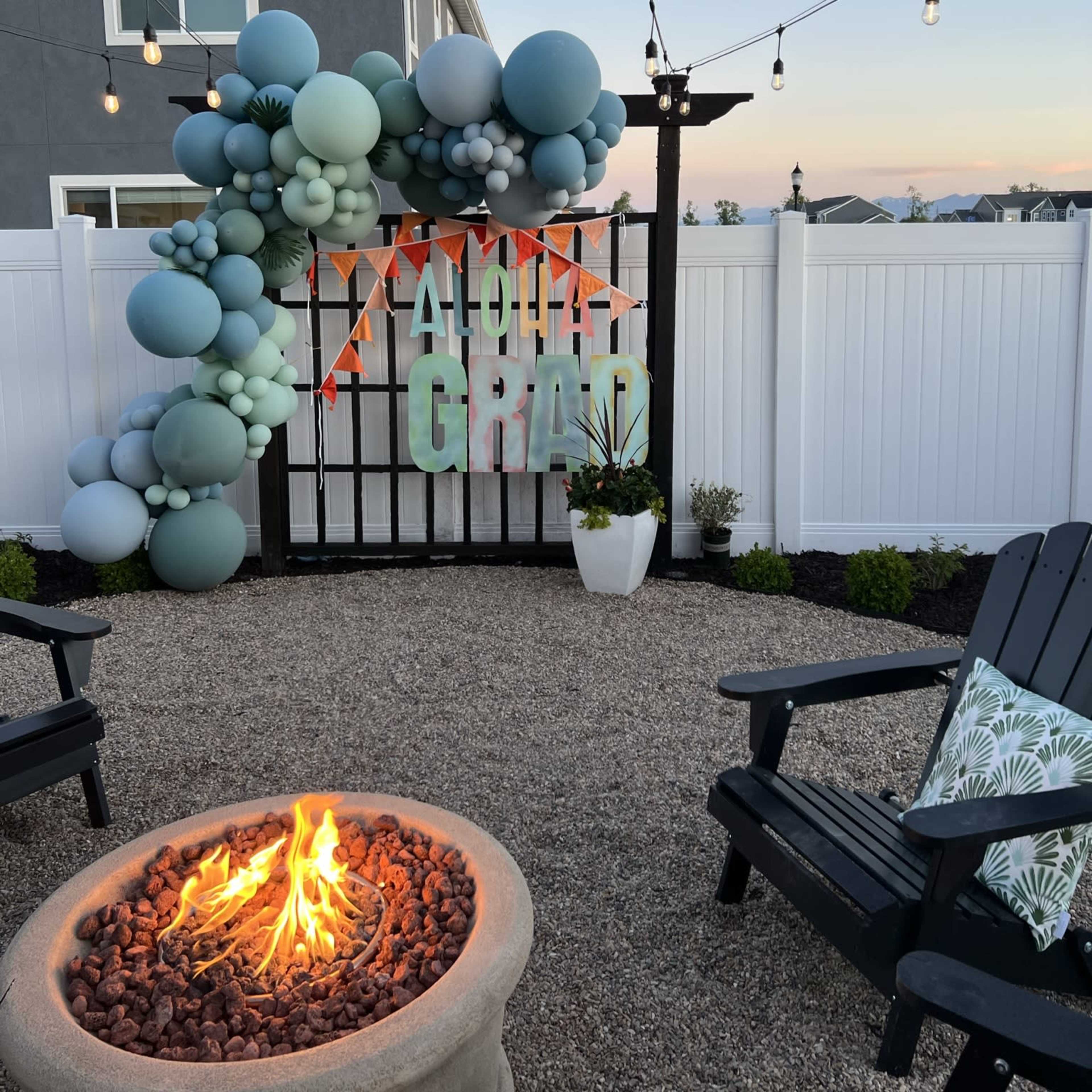 A decorative graduation backdrop featuring balloons in various shades of blue and green is set up in a gravel patio area with string lights and a fire pit.
