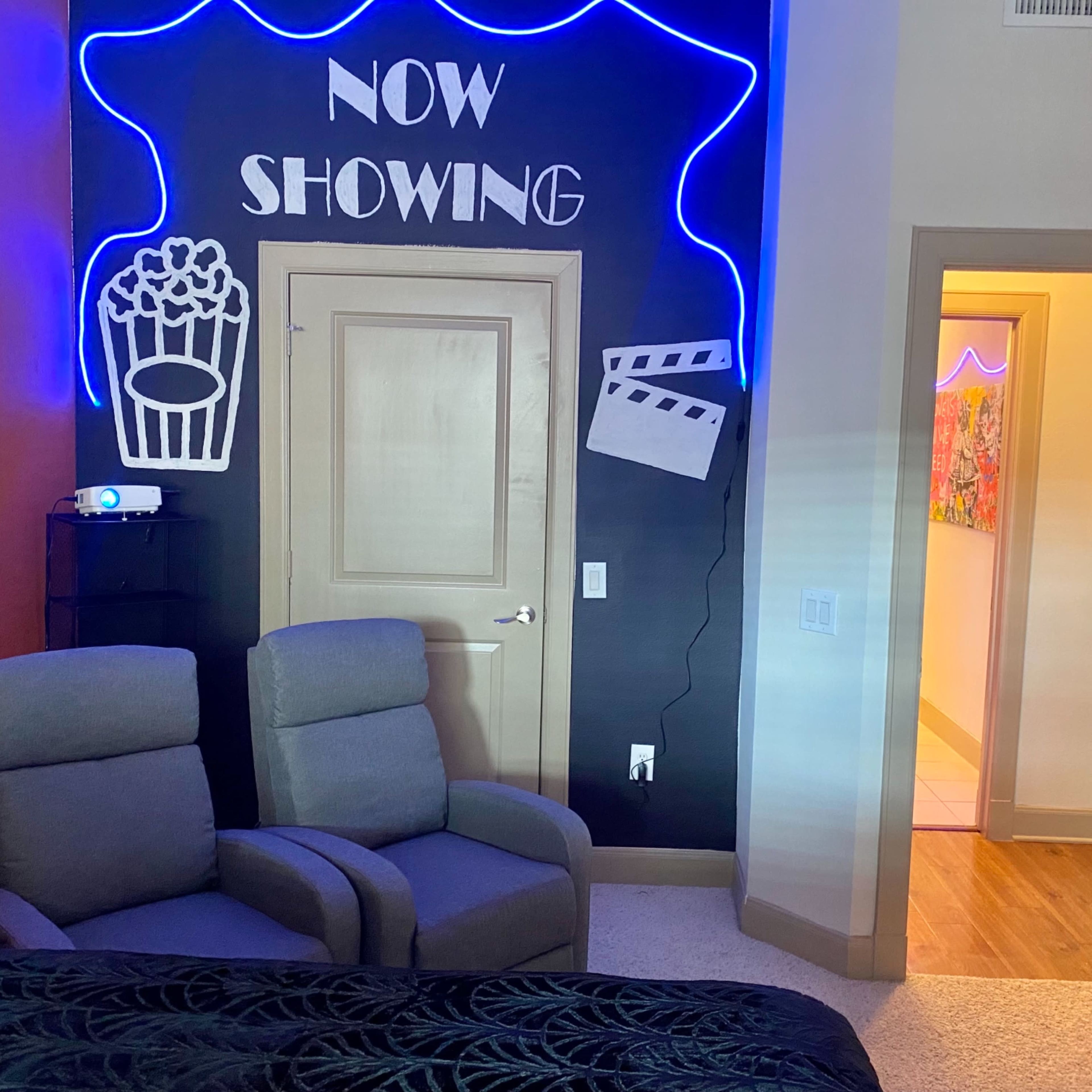 A cozy room with a projector setup, featuring a "Now Showing" neon sign, movie-themed decorations, and two recliner chairs.