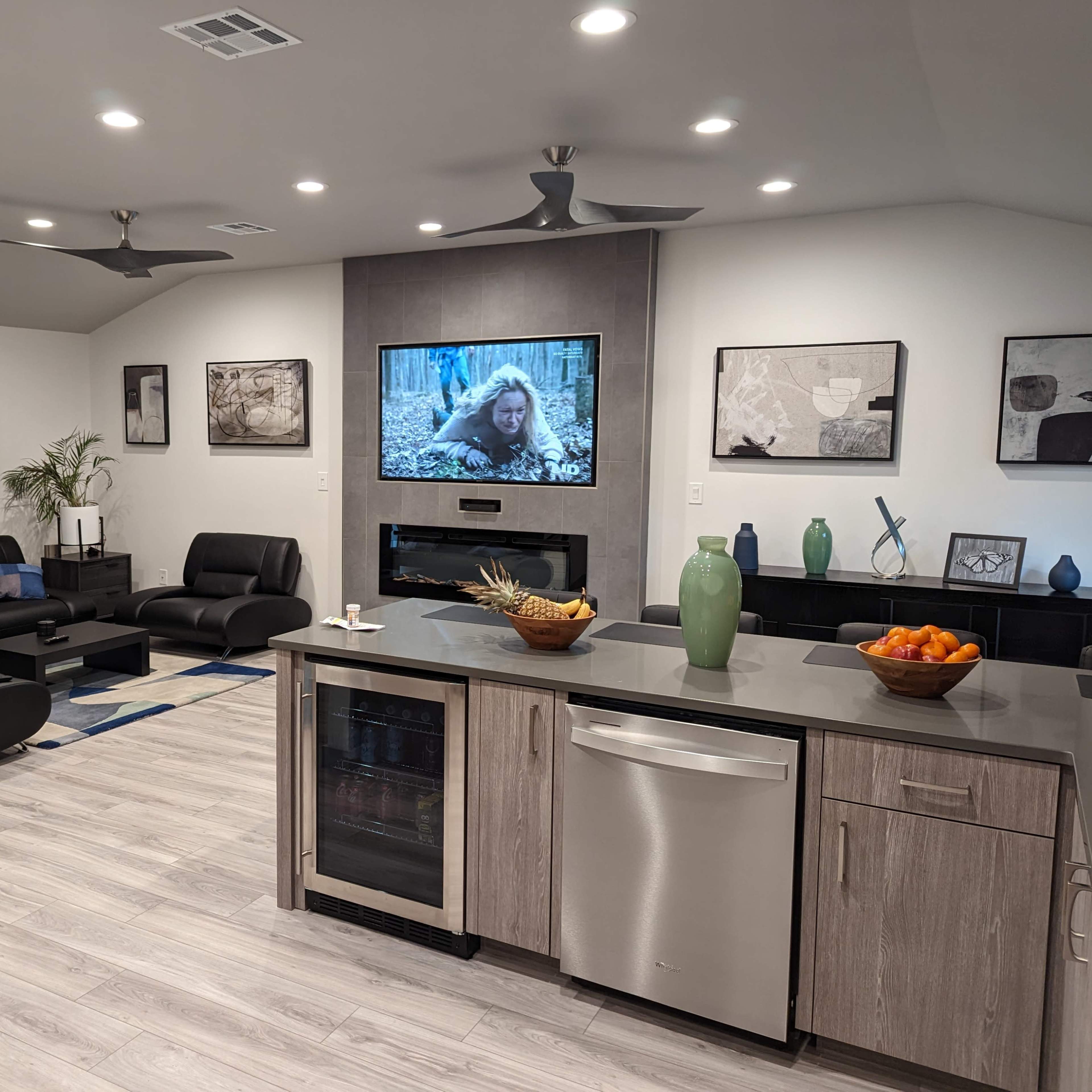 A modern living room and kitchen area featuring dark furniture, a large wall-mounted television displaying a scene from a movie, and a kitchen island with various decorative items and fruit.