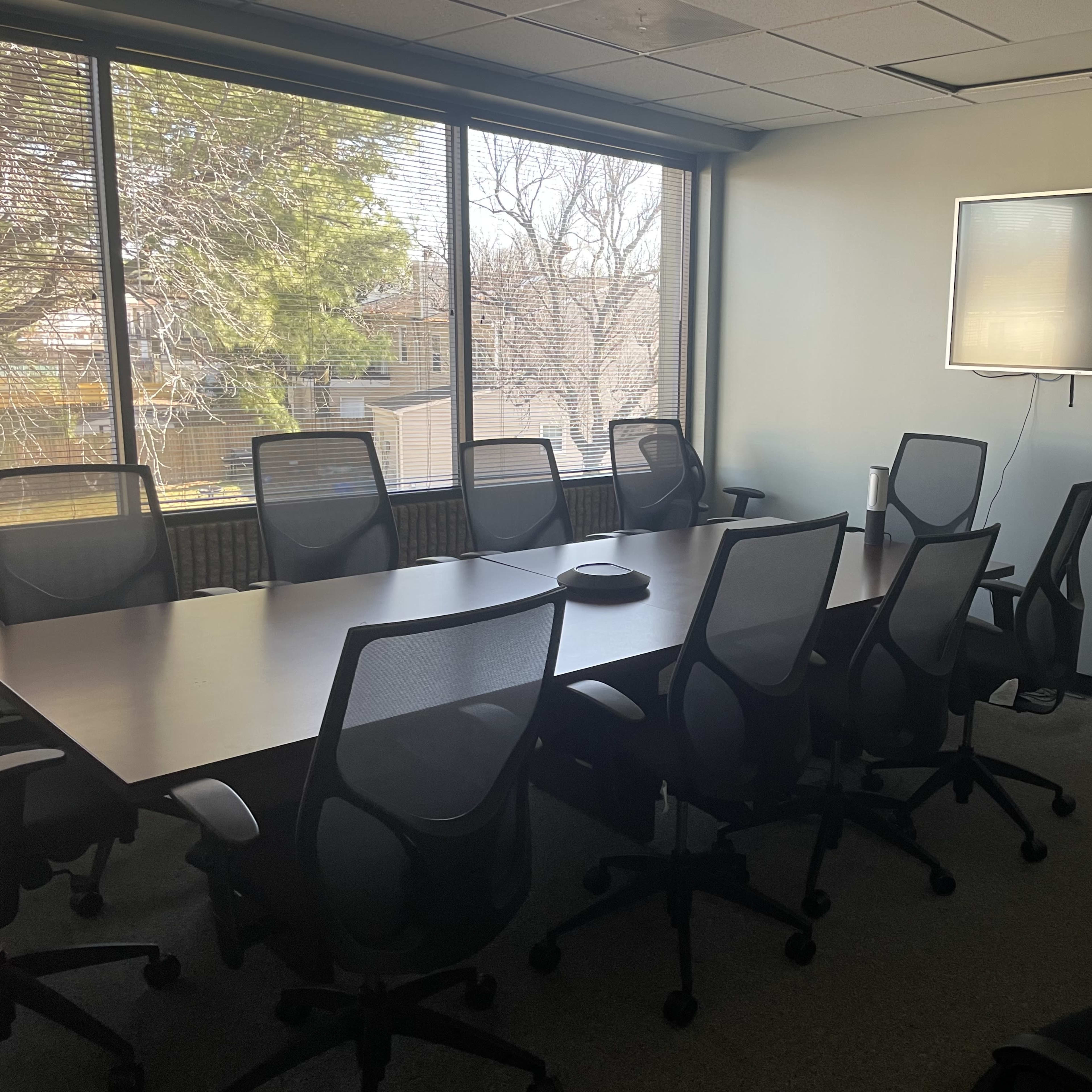 A meeting room with a large conference table surrounded by eight chairs, a wall-mounted television, and a whiteboard, all set against a backdrop of large windows.