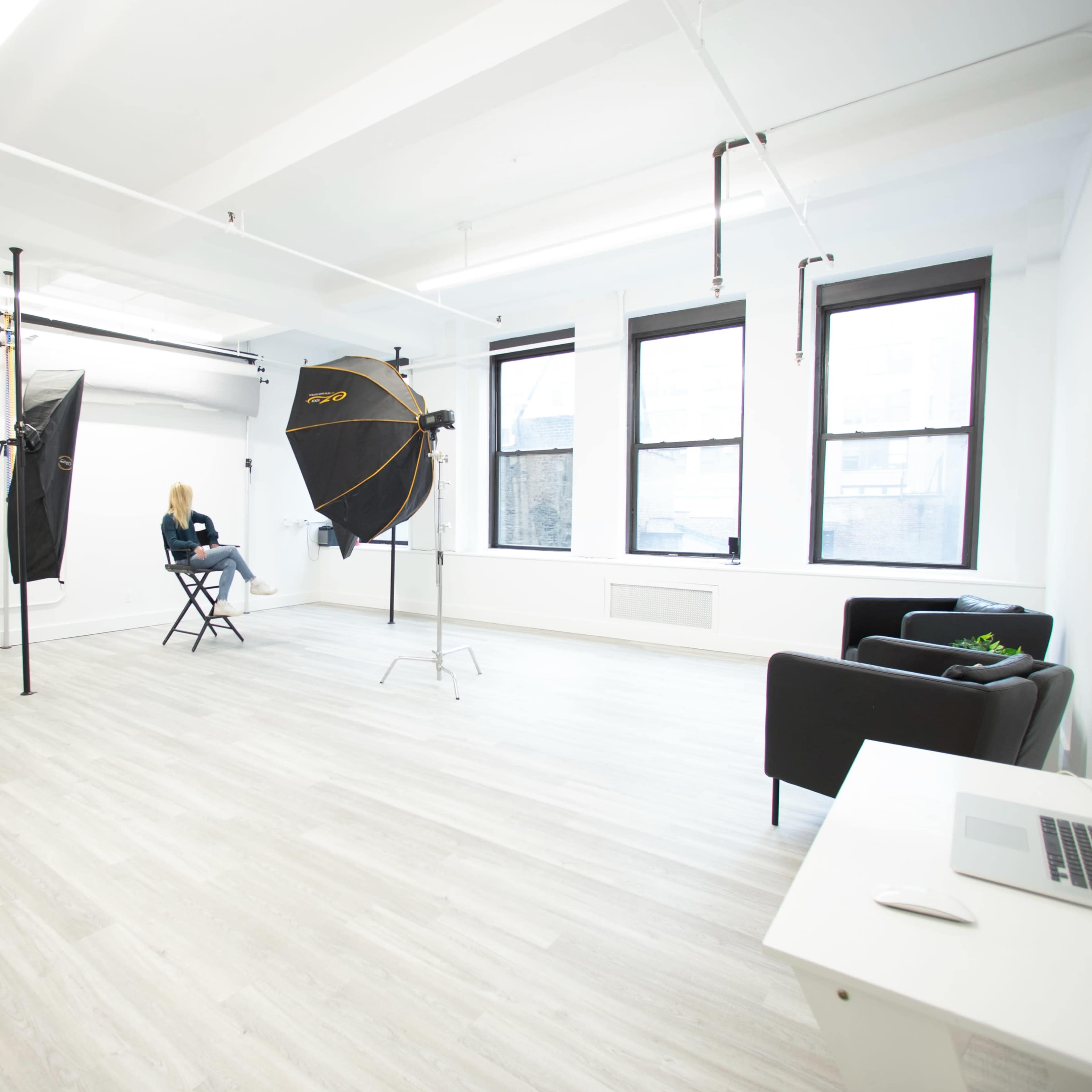 A bright photography studio with two softbox lights, a vacant chair, and a small seating area.