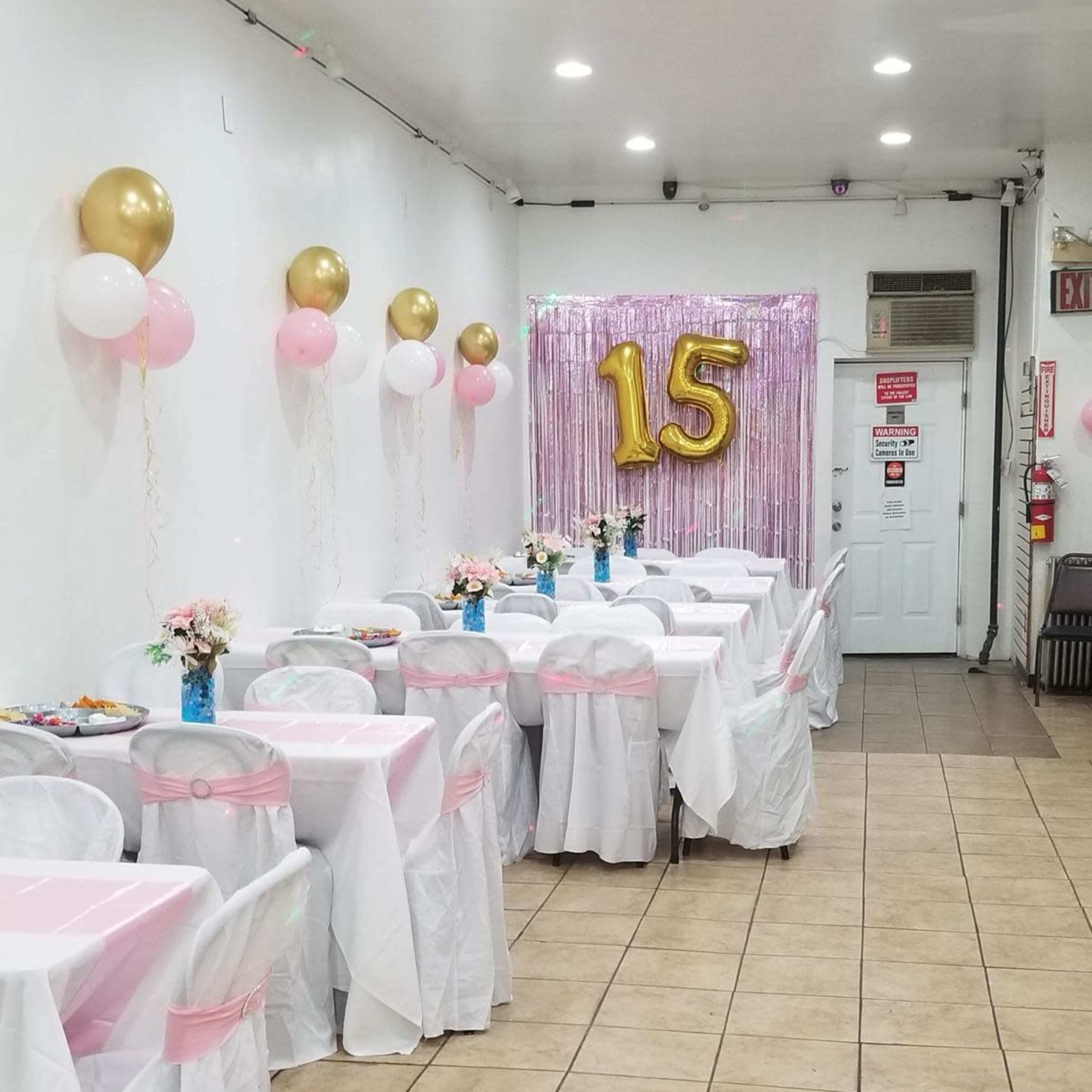 The room is set up for a celebration with tables dressed in white tablecloths and pink accents, adorned with flowers, balloons, and a large golden "15" decoration on the wall.