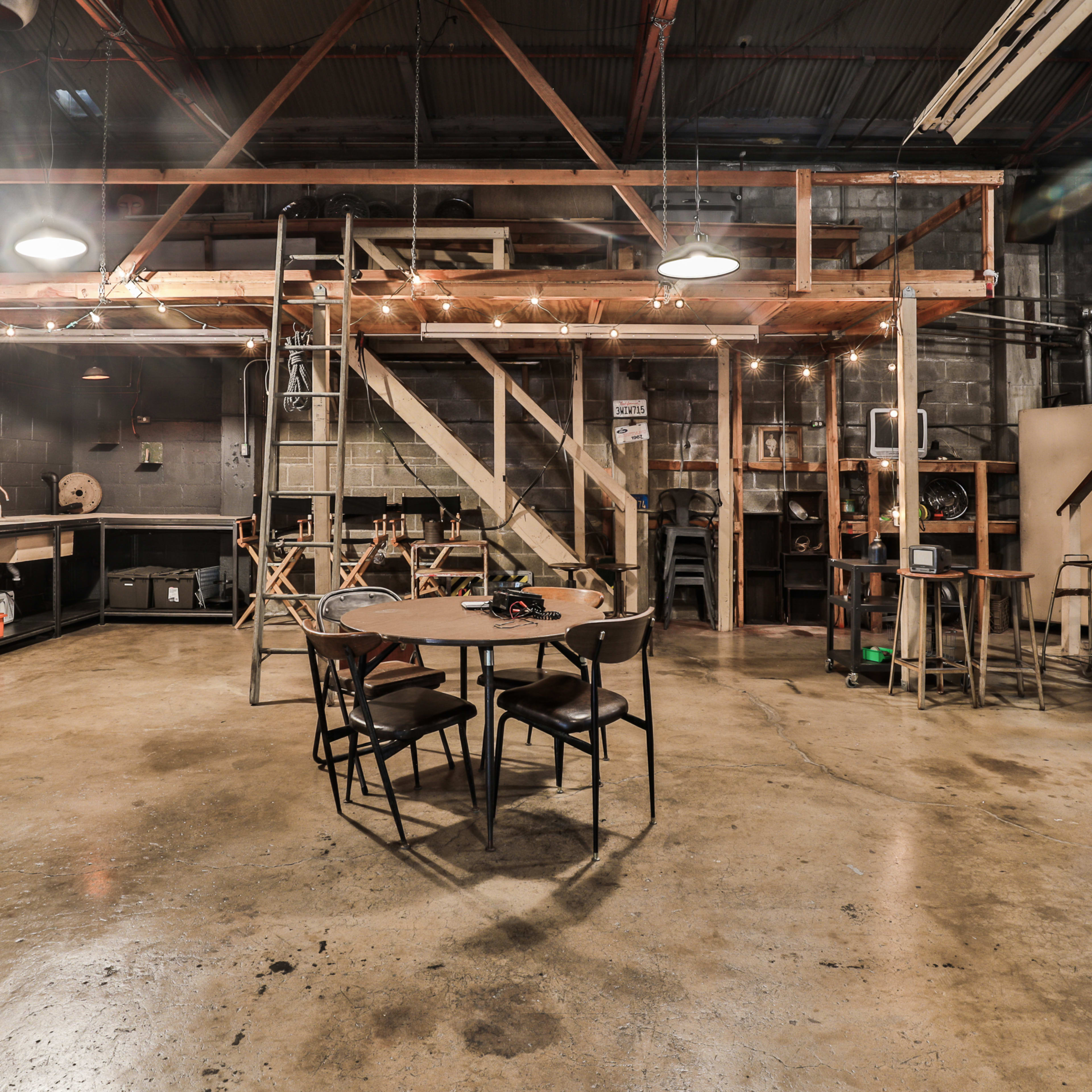 An industrial-style workshop featuring a wooden loft, a central table with chairs, and various tools and equipment along the walls.