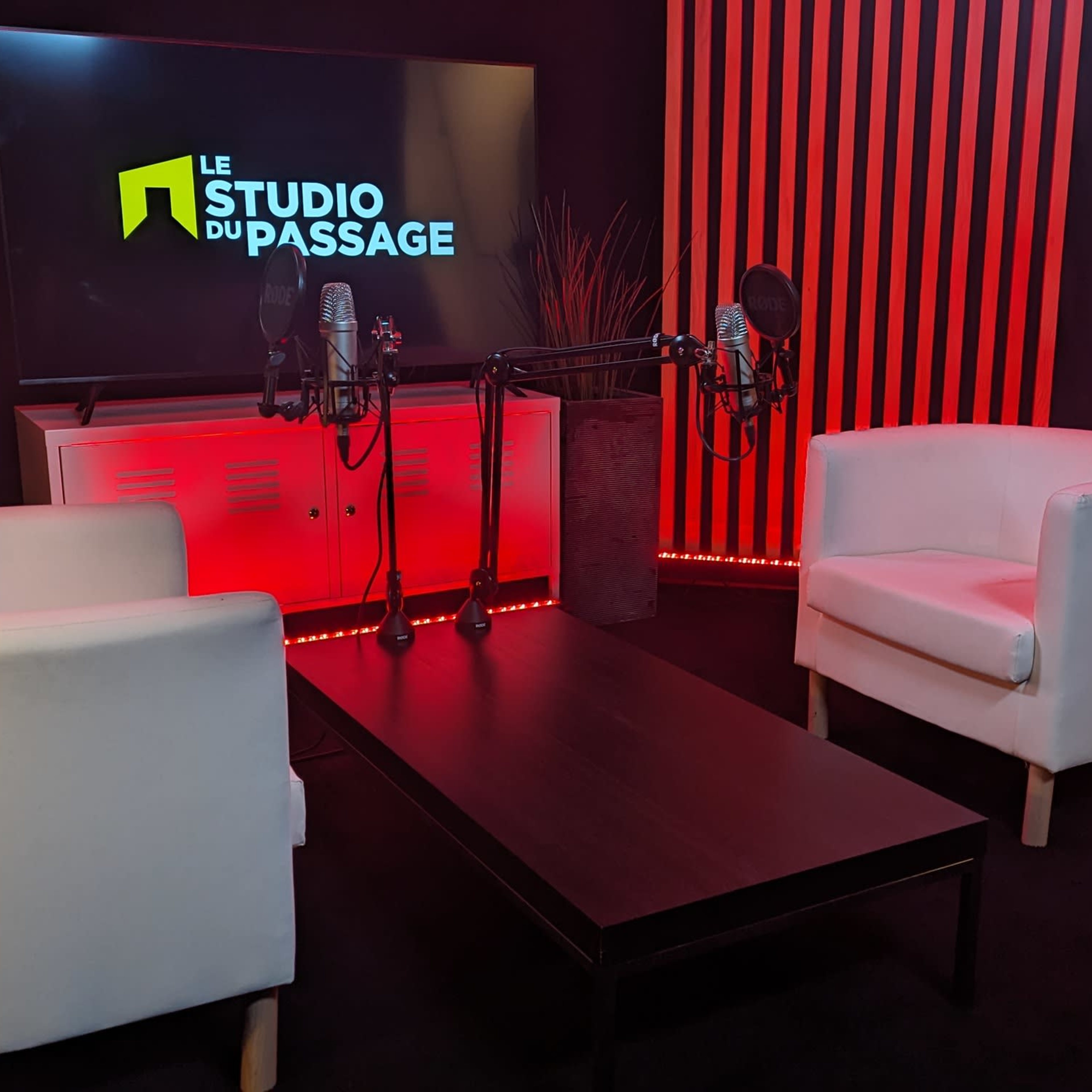 A modern studio setup with two white chairs, a black table, and two microphones in front of a screen displaying "LE STUDIO DU PASSAGE" against a backdrop of red and black stripes.