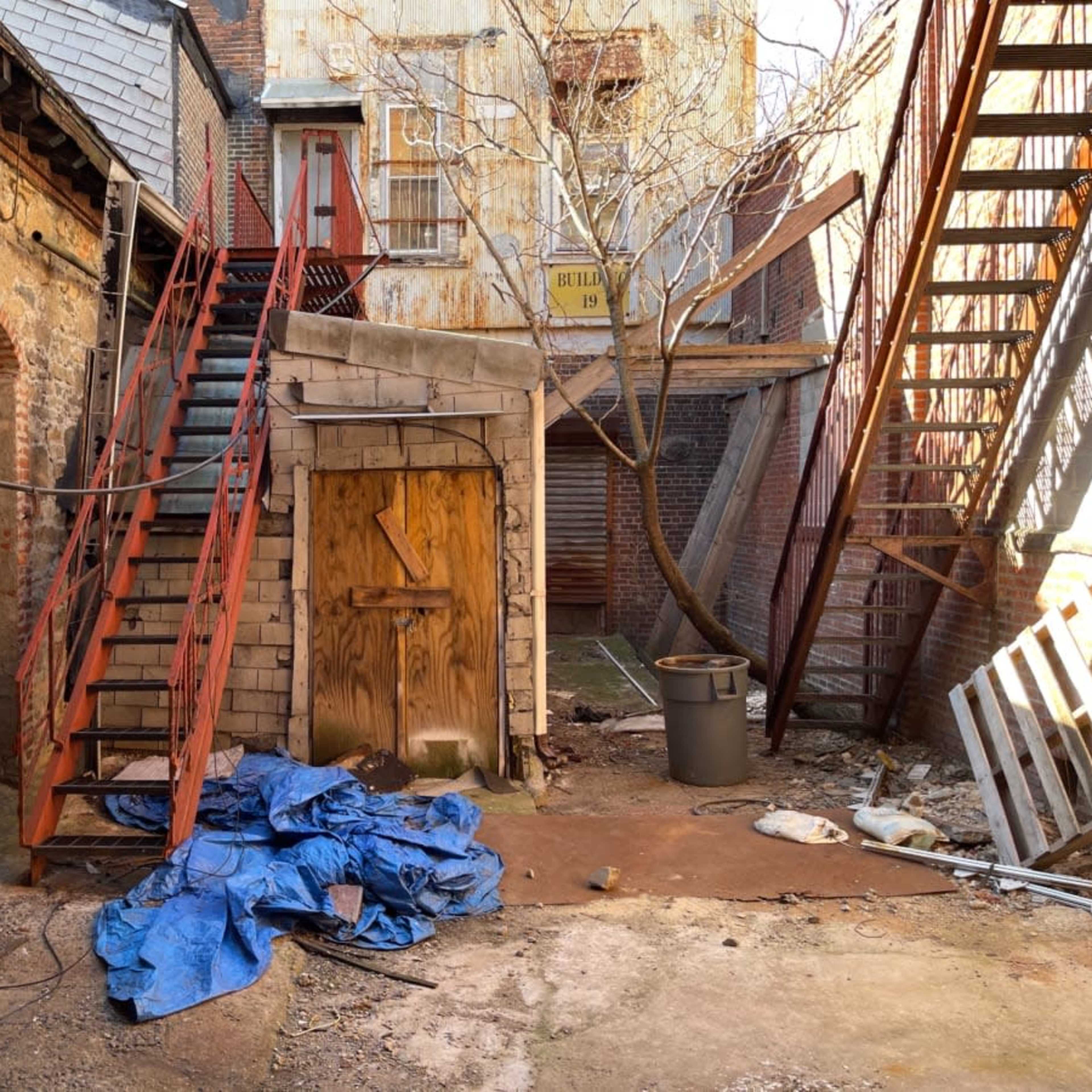 A rundown alleyway featuring a wooden door, a blue tarp on the ground, and a staircase leading to a second level.