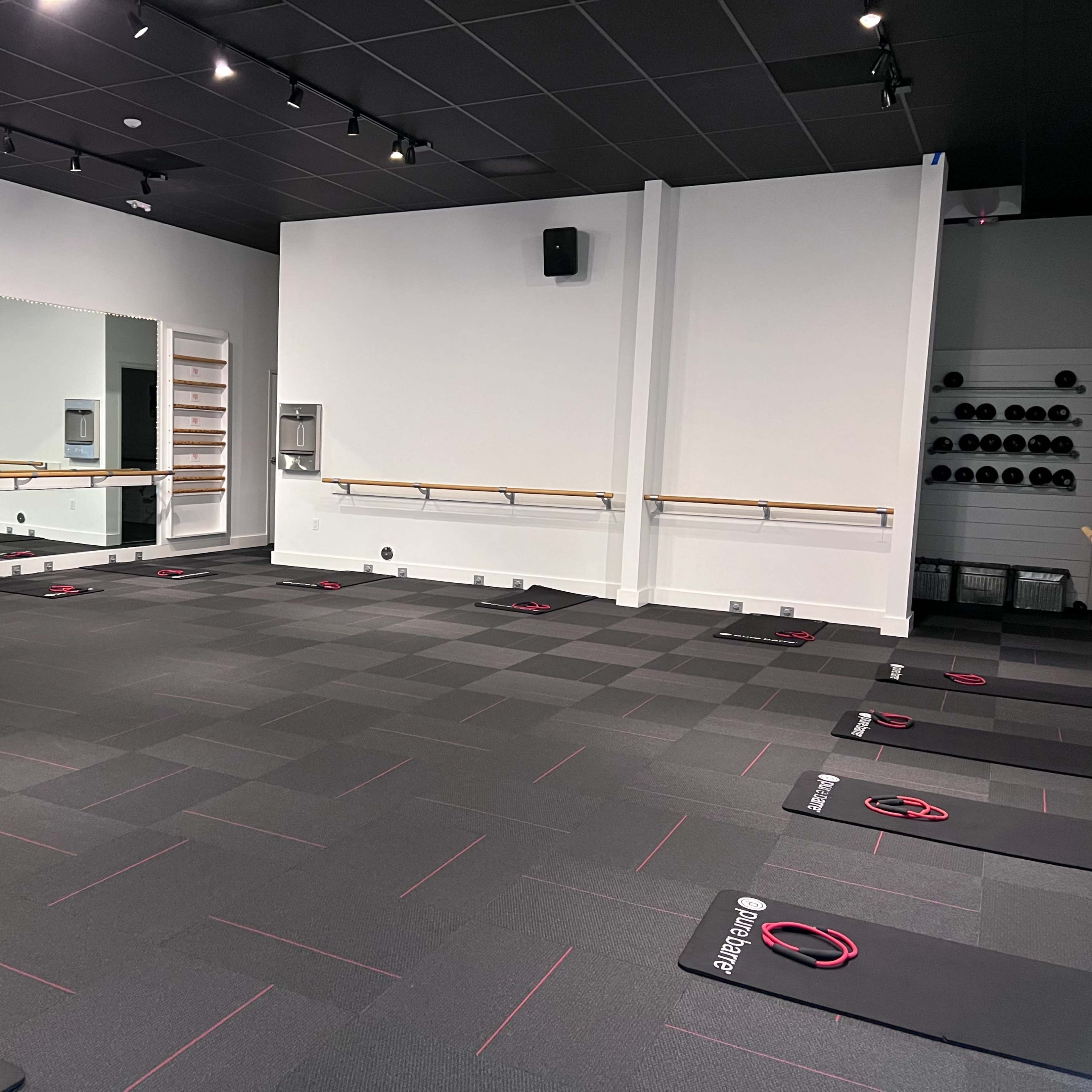 A fitness studio with exercise mats arranged on the floor, a large mirror on one wall, and a row of weights on shelves.