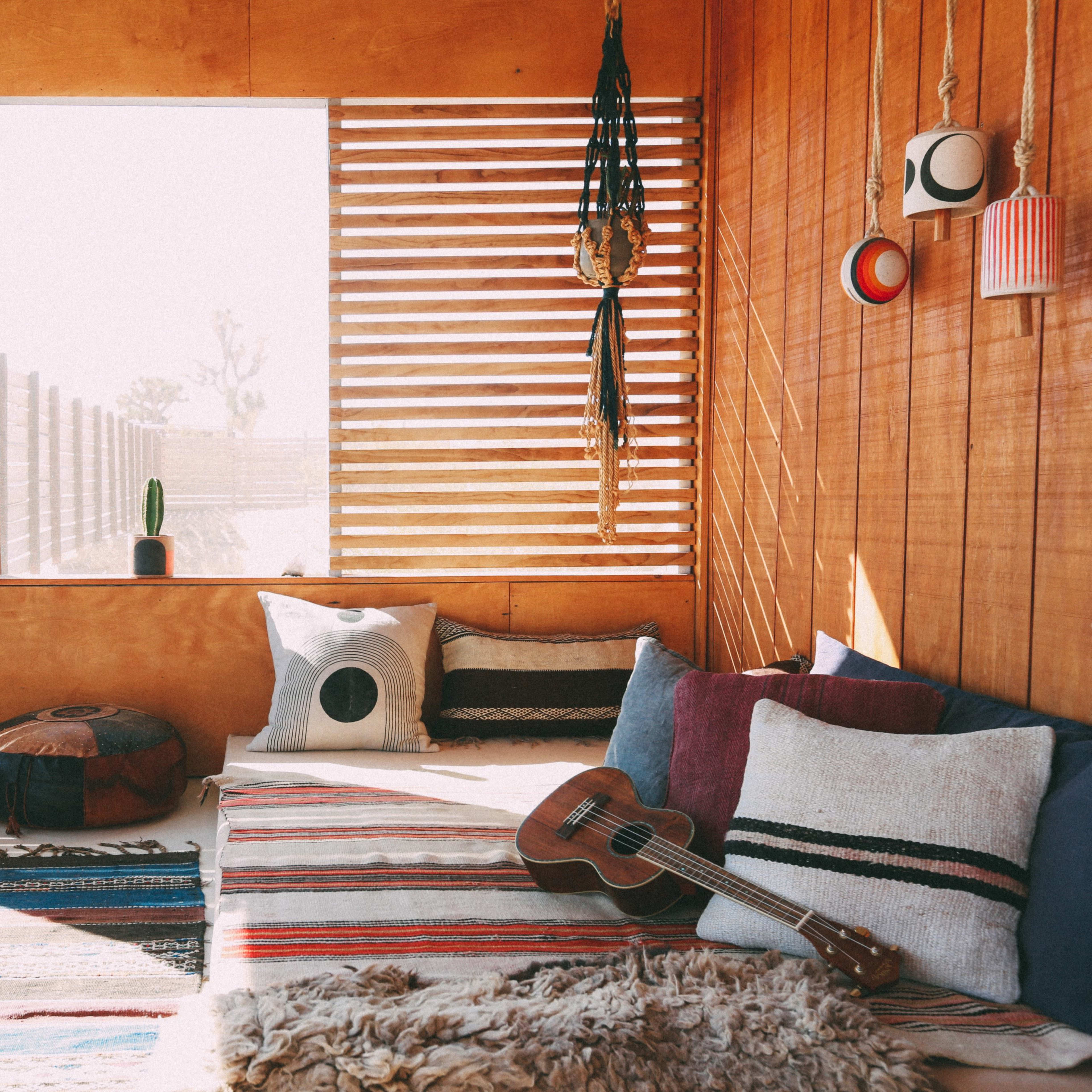 The scene features a cozy interior with wooden walls, a seating area adorned with patterned rugs and pillows, a ukulele resting nearby, and large windows allowing natural light to enter.