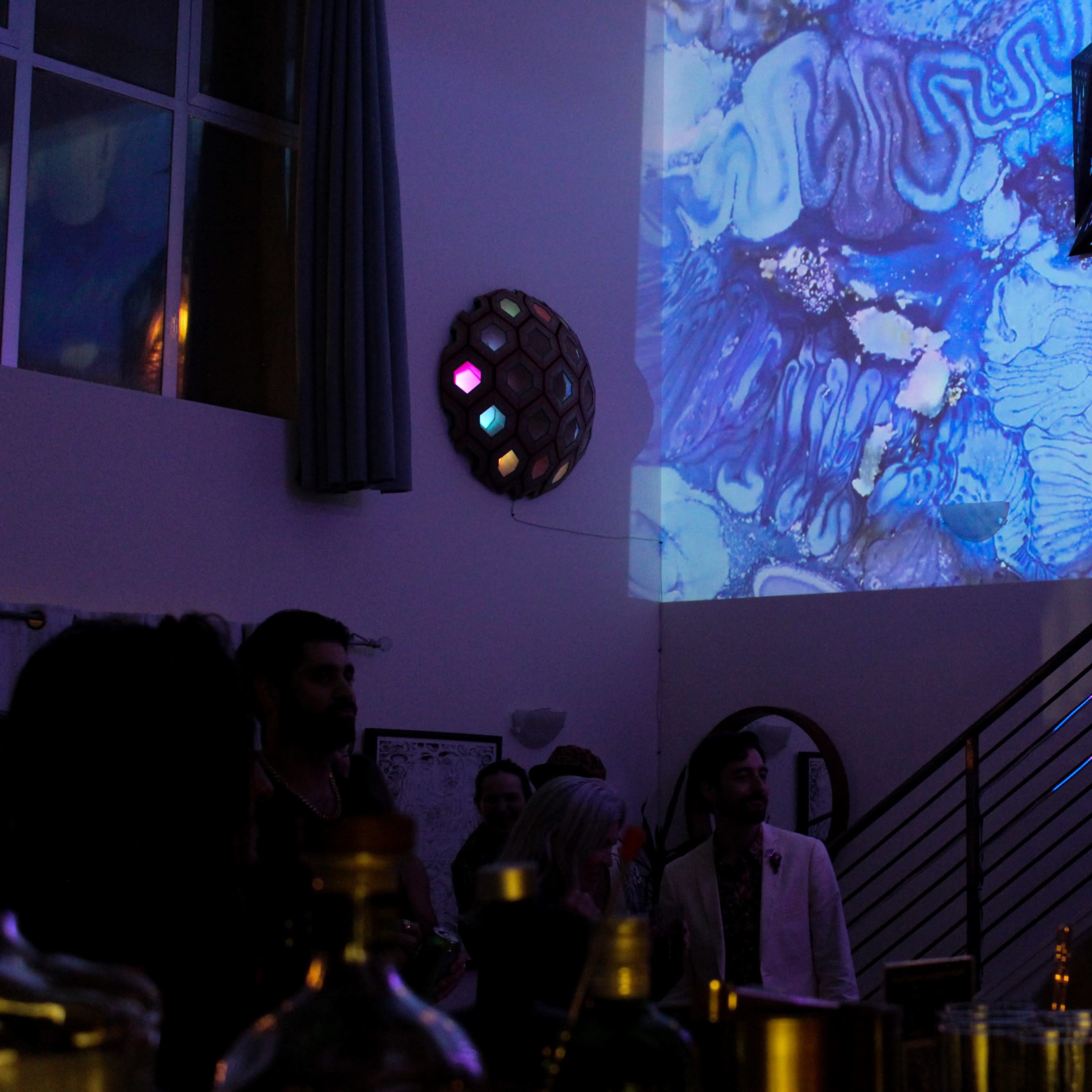 A geometric lamp hangs from the ceiling while a colorful projection illuminates a wall in an indoor social setting.