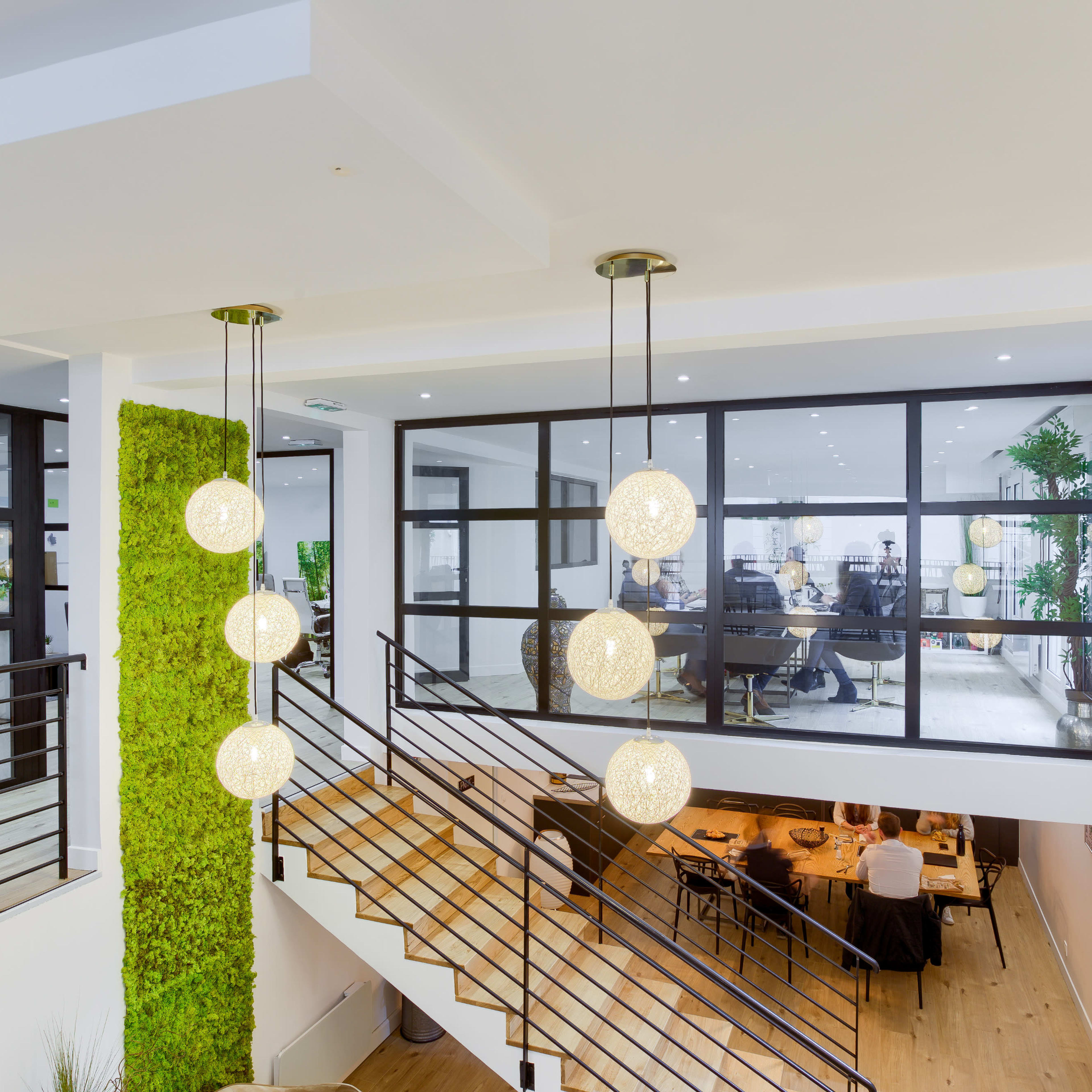 A modern interior features a staircase with hanging spherical lights, surrounded by greenery and large windows overlooking a dining area.