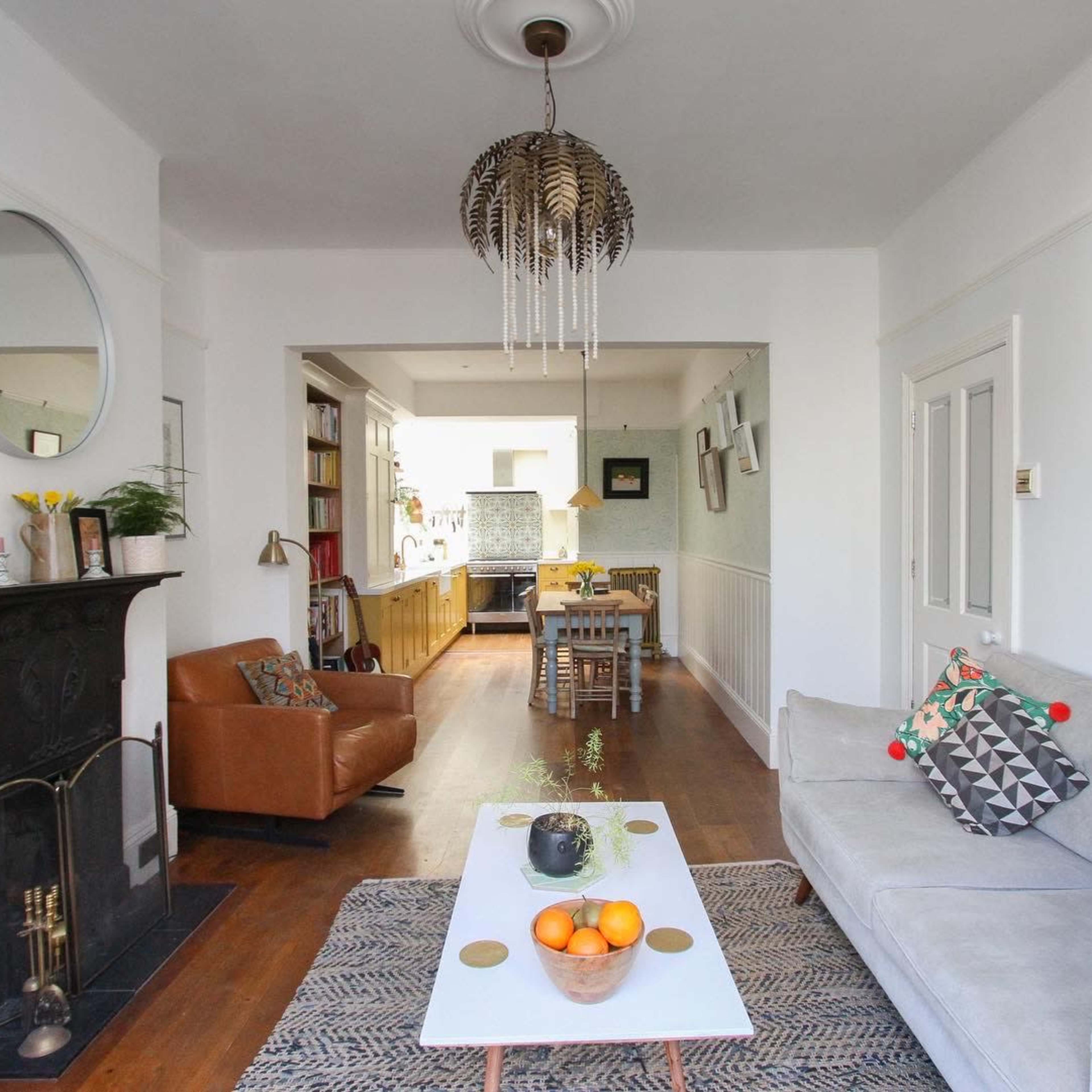 A living room features a sofa, coffee table with oranges, a fireplace, and an adjoining kitchen, all set against light-colored walls and wooden floors.