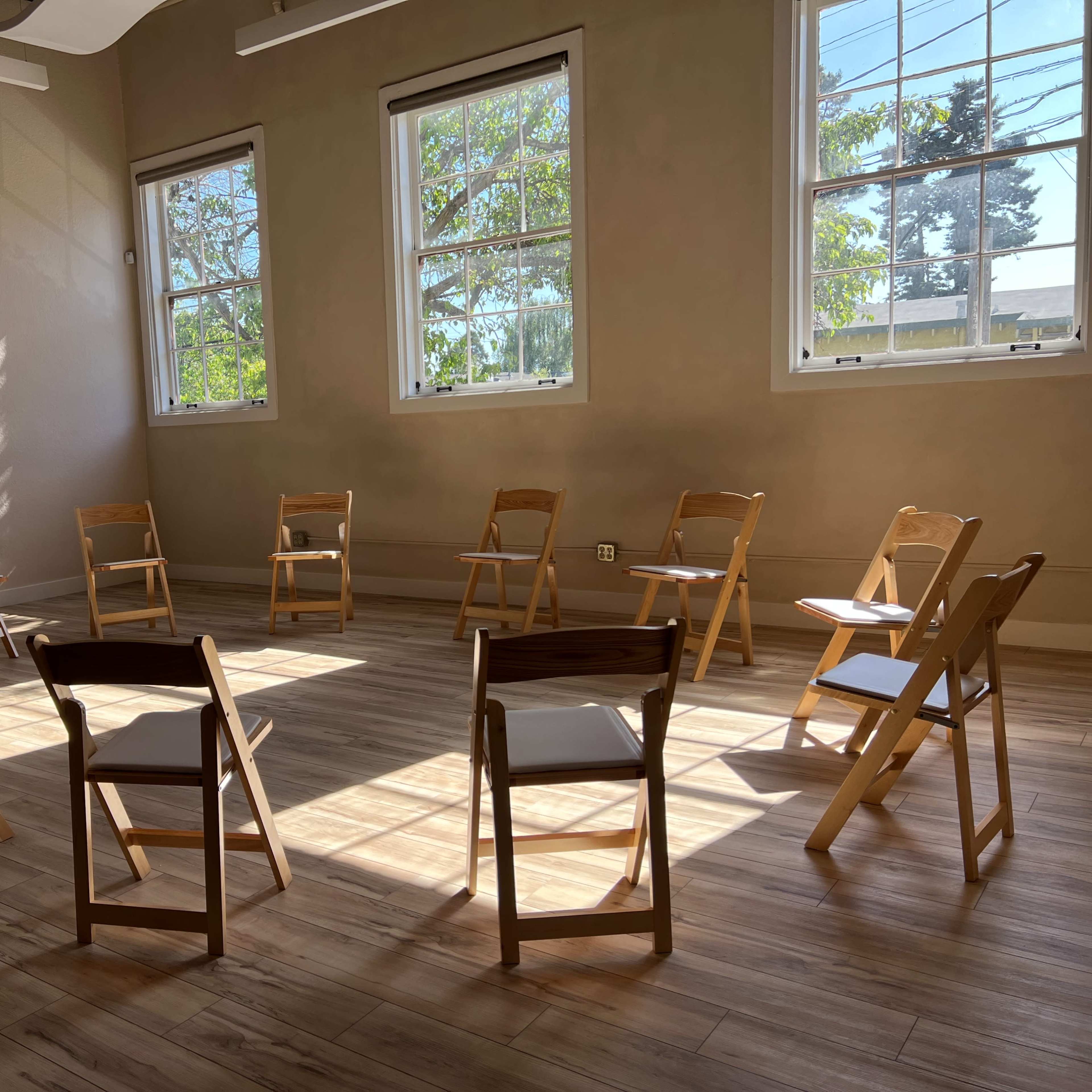 A circle of wooden chairs is arranged in a sunlit room with large windows and light-colored walls.