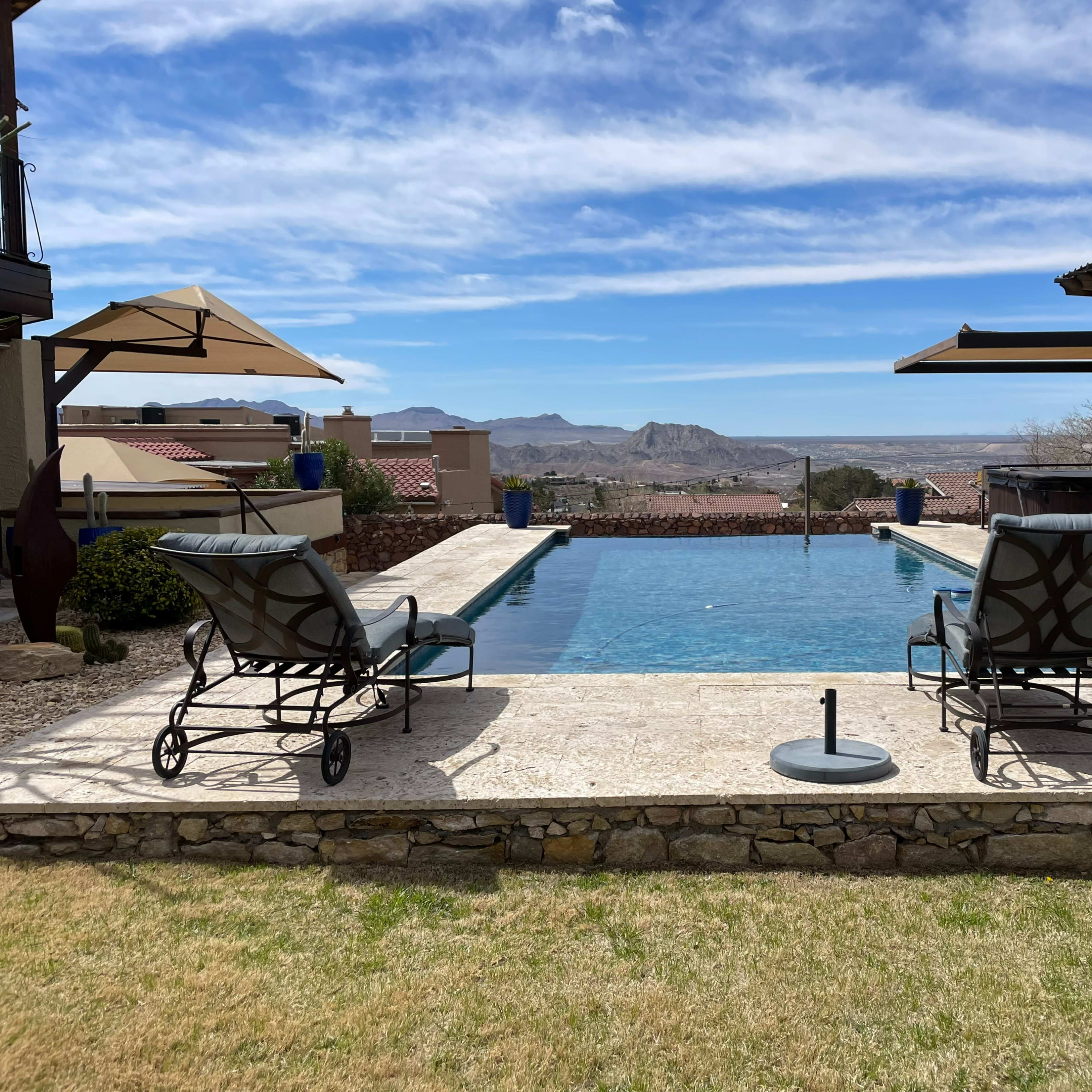 A swimming pool with two lounge chairs beside it, overlooking a mountainous landscape under a partly cloudy sky.