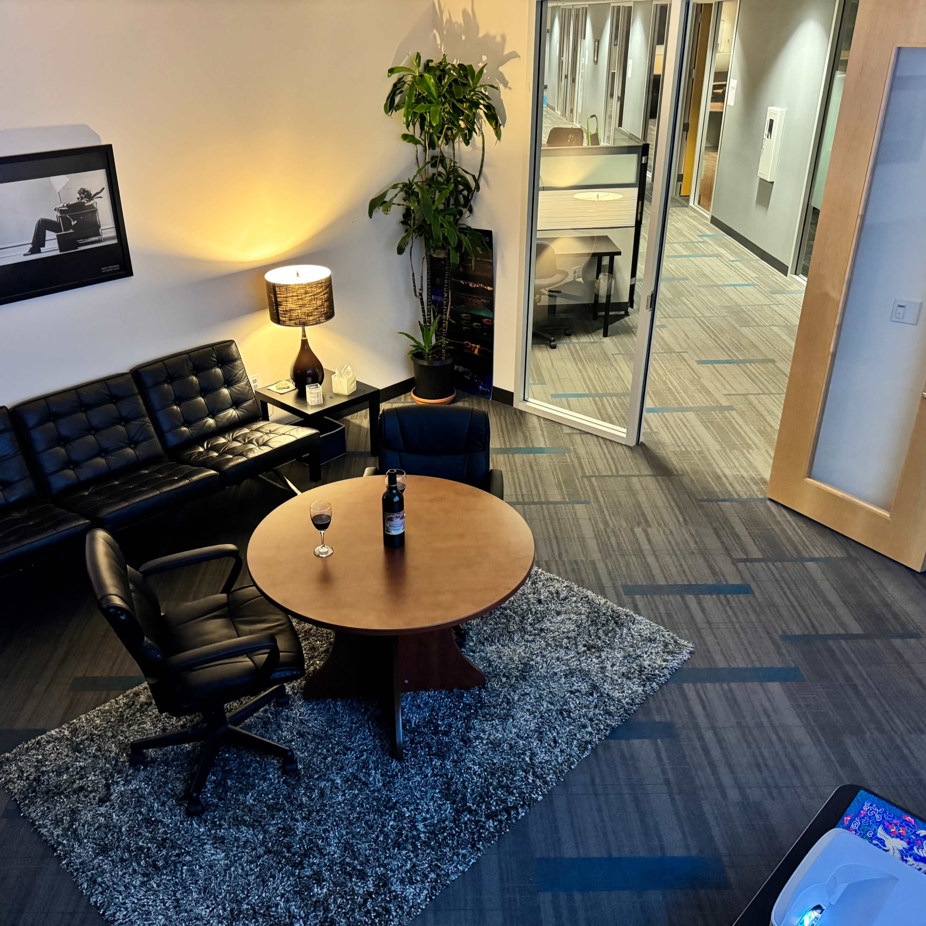 A modern office lounge area featuring a round wooden table, leather chairs, a sofa, and a glass of wine on the table, with a plant in the corner and an open doorway leading to another workspace.