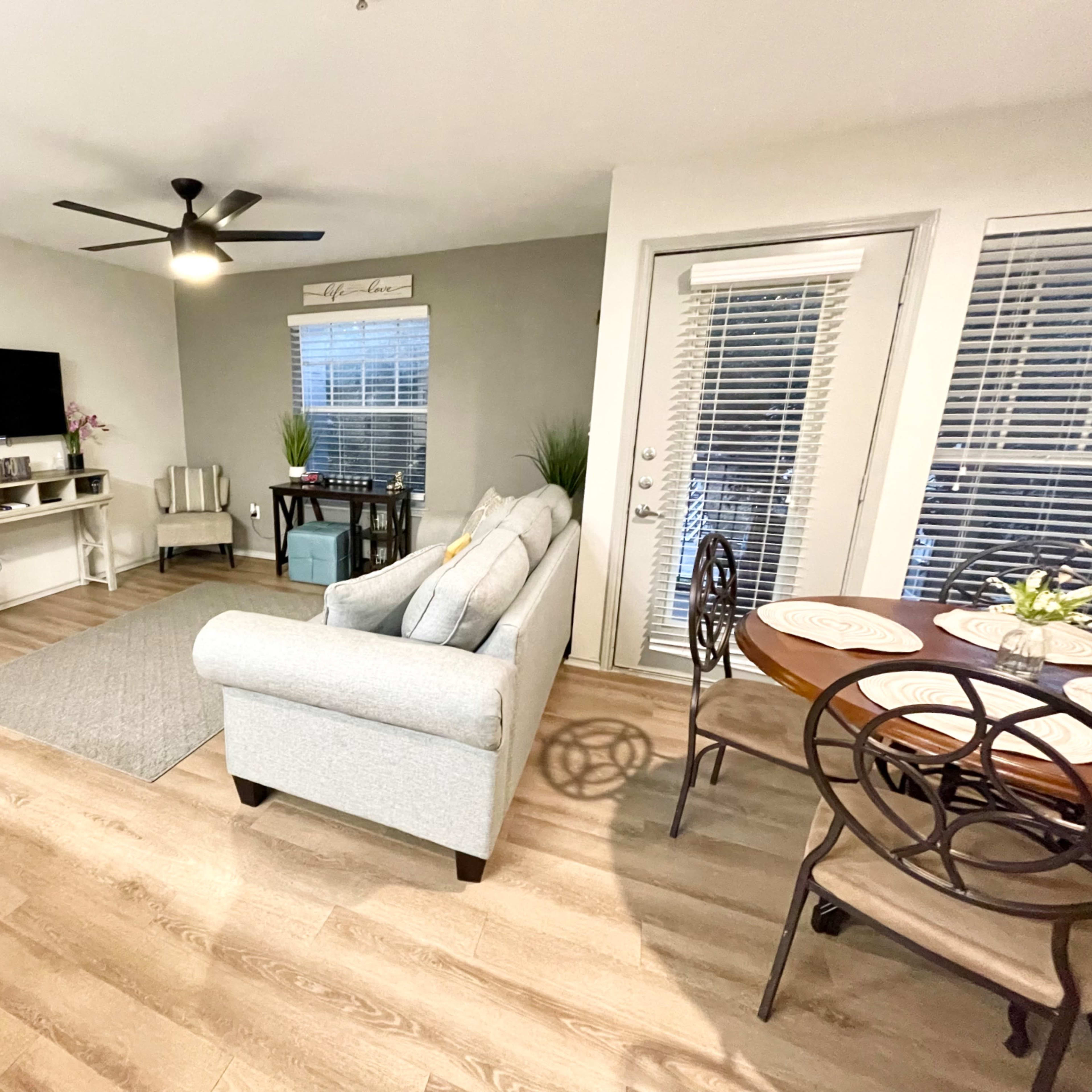 A living room with a light-colored sofa, a small dining table by the window, and a television mounted on the wall.