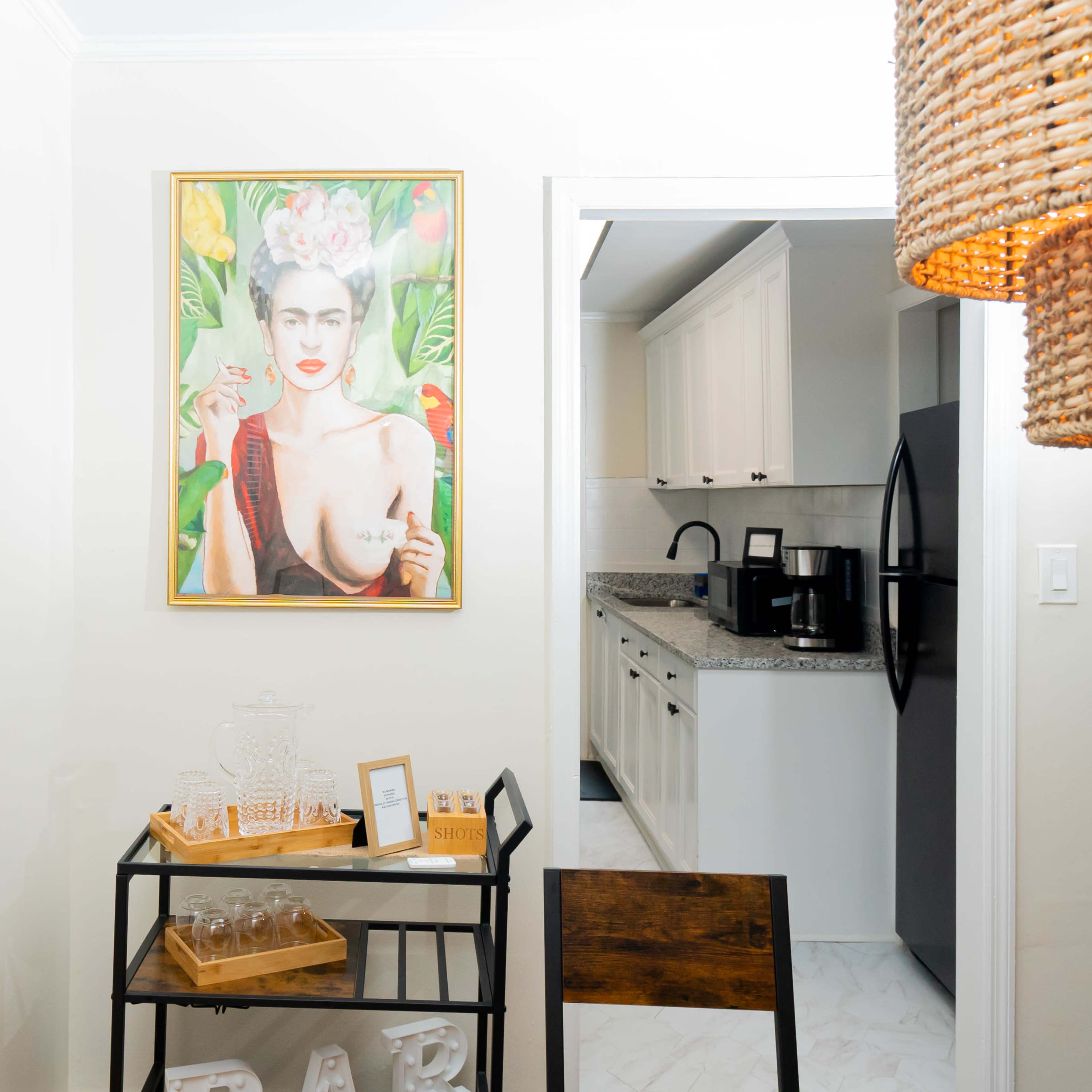A framed portrait of a woman with flowers in her hair hangs on a light wall opposite a kitchen area with black appliances and white cabinets.