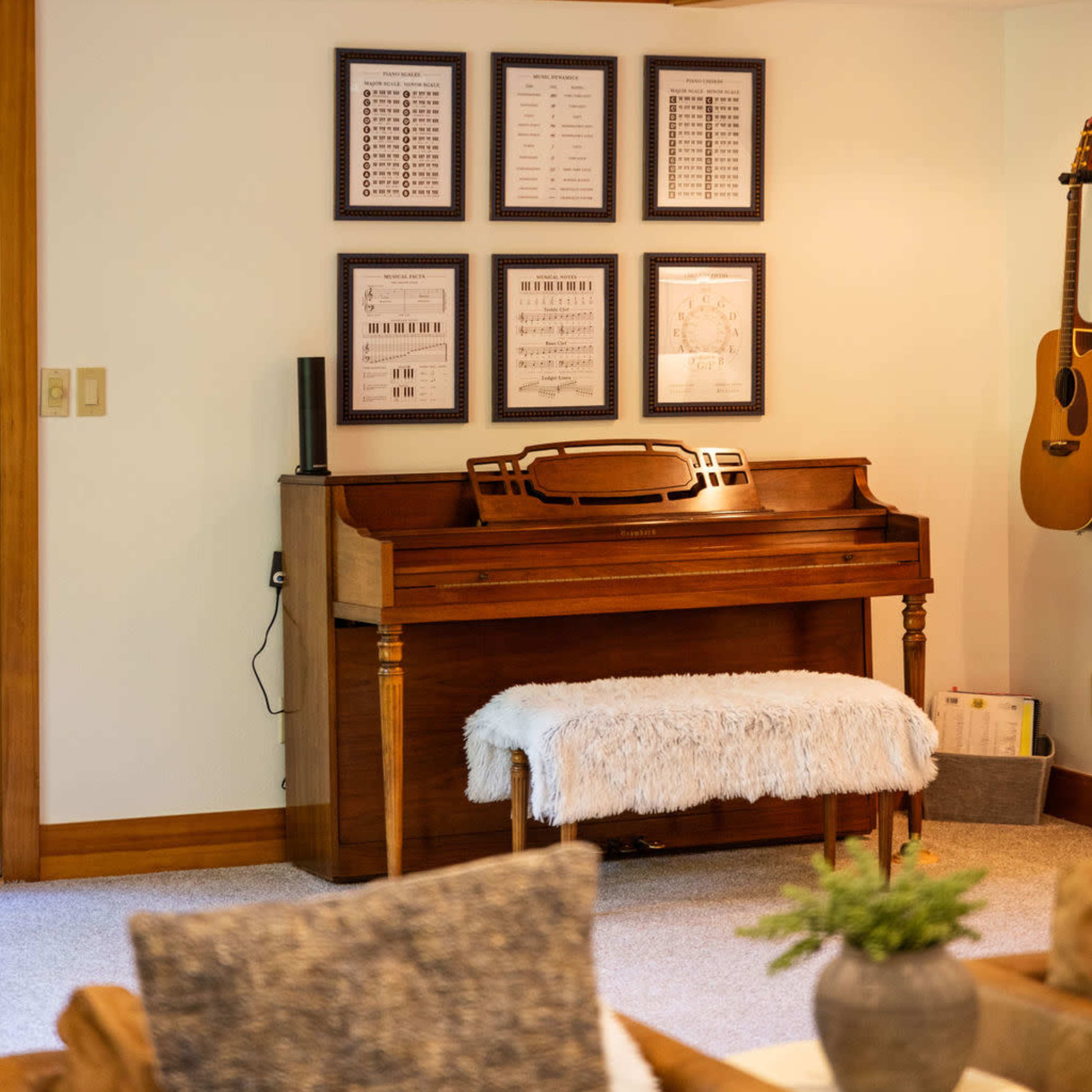 A cozy room with a wooden piano against the wall, decorated with framed musical sheets, and a guitar hanging nearby.