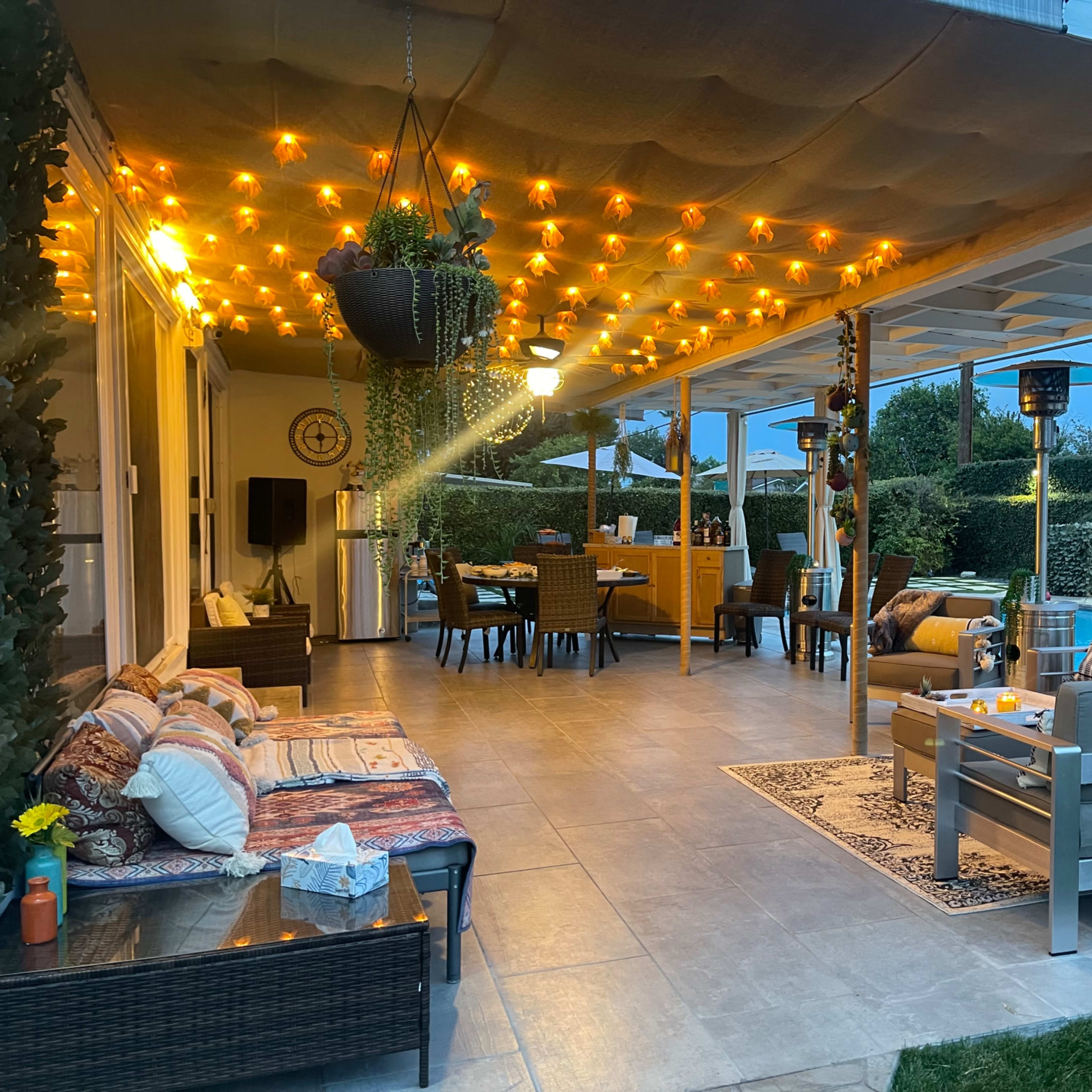A covered outdoor patio features stylish seating, warm string lights, and a dining area surrounded by lush greenery.