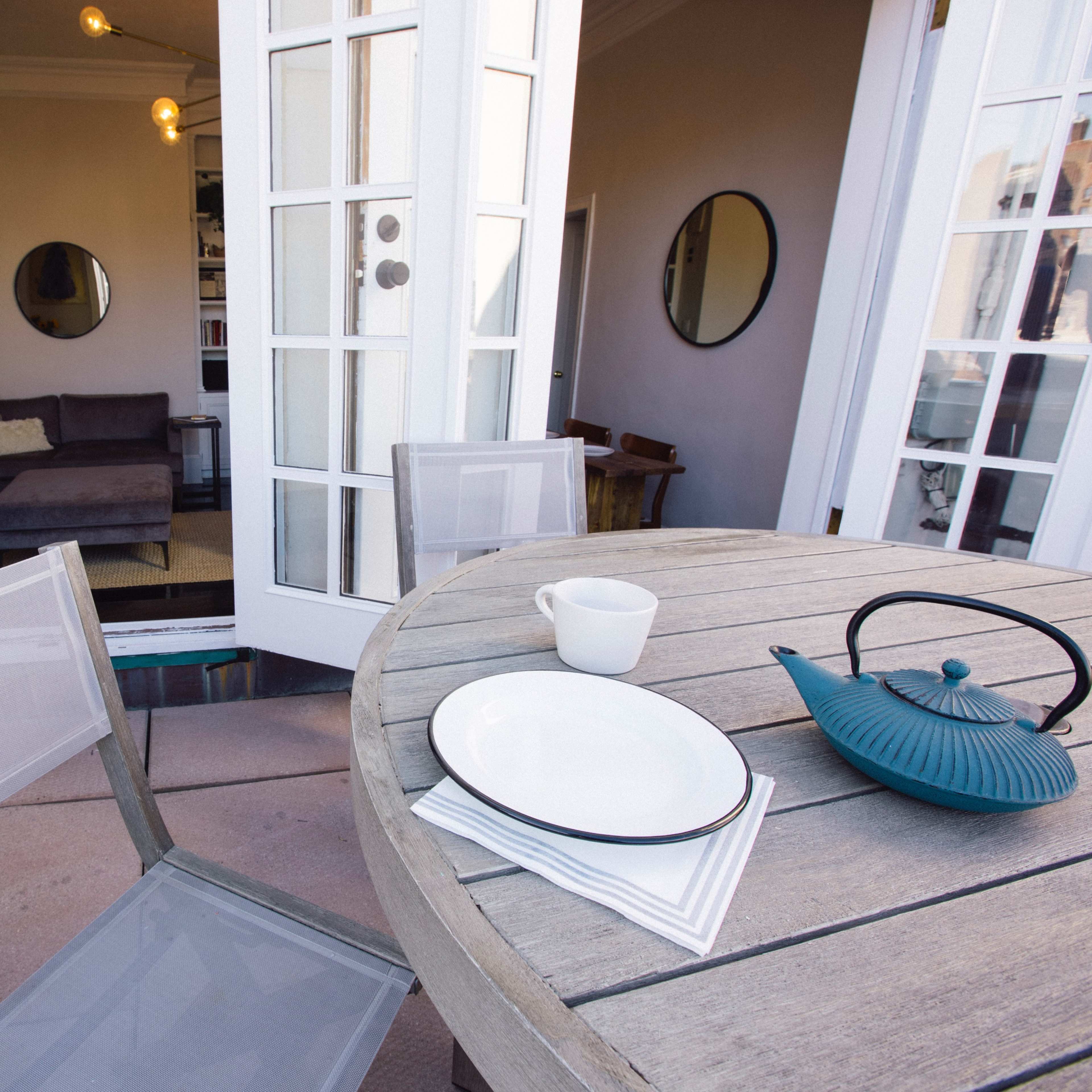 A round wooden table with a teapot and plate sits on a balcony overlooking an indoor living space with large windows.