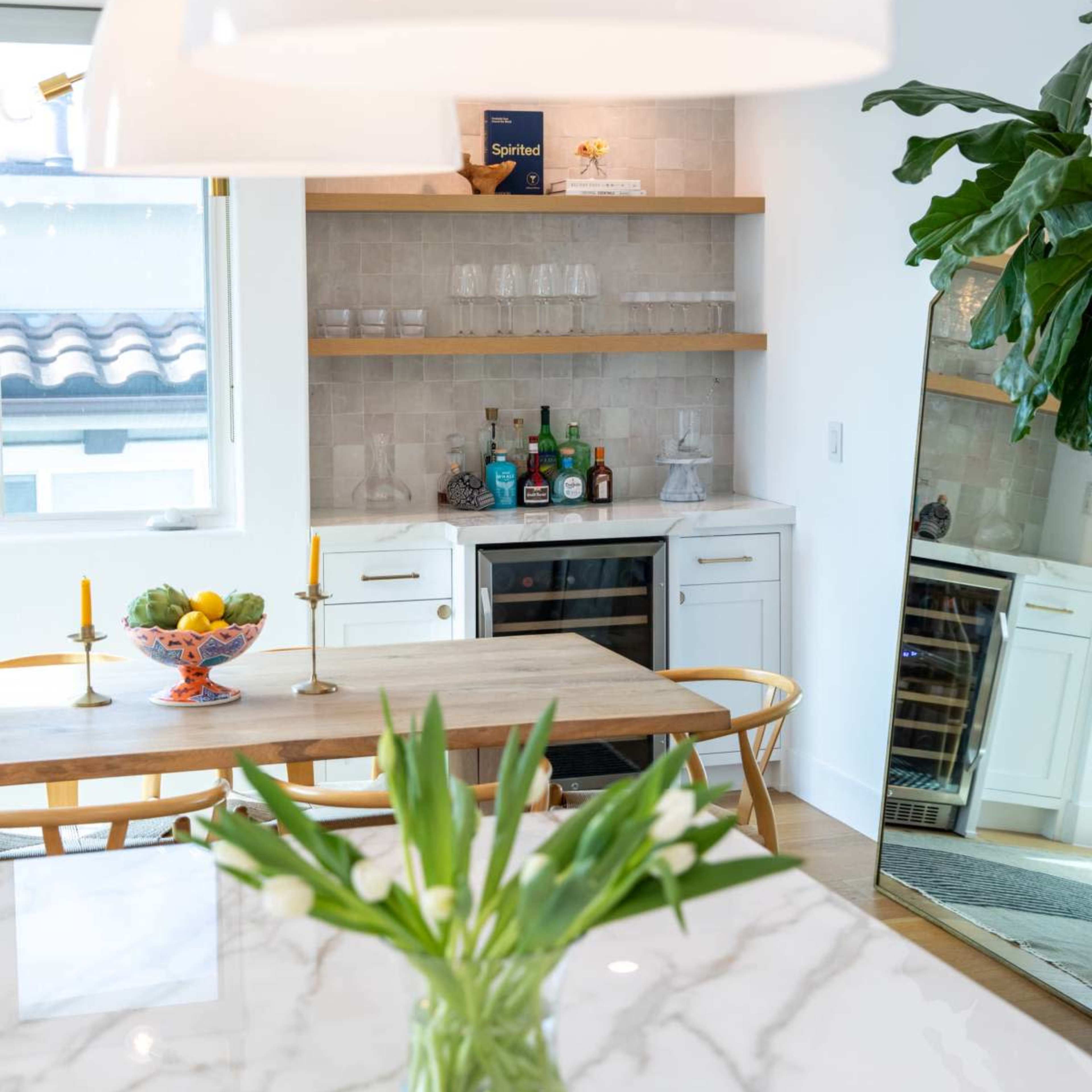 A modern kitchen features a dining table with fruit, a bar area with assorted drinks, and a large mirror reflecting the space.