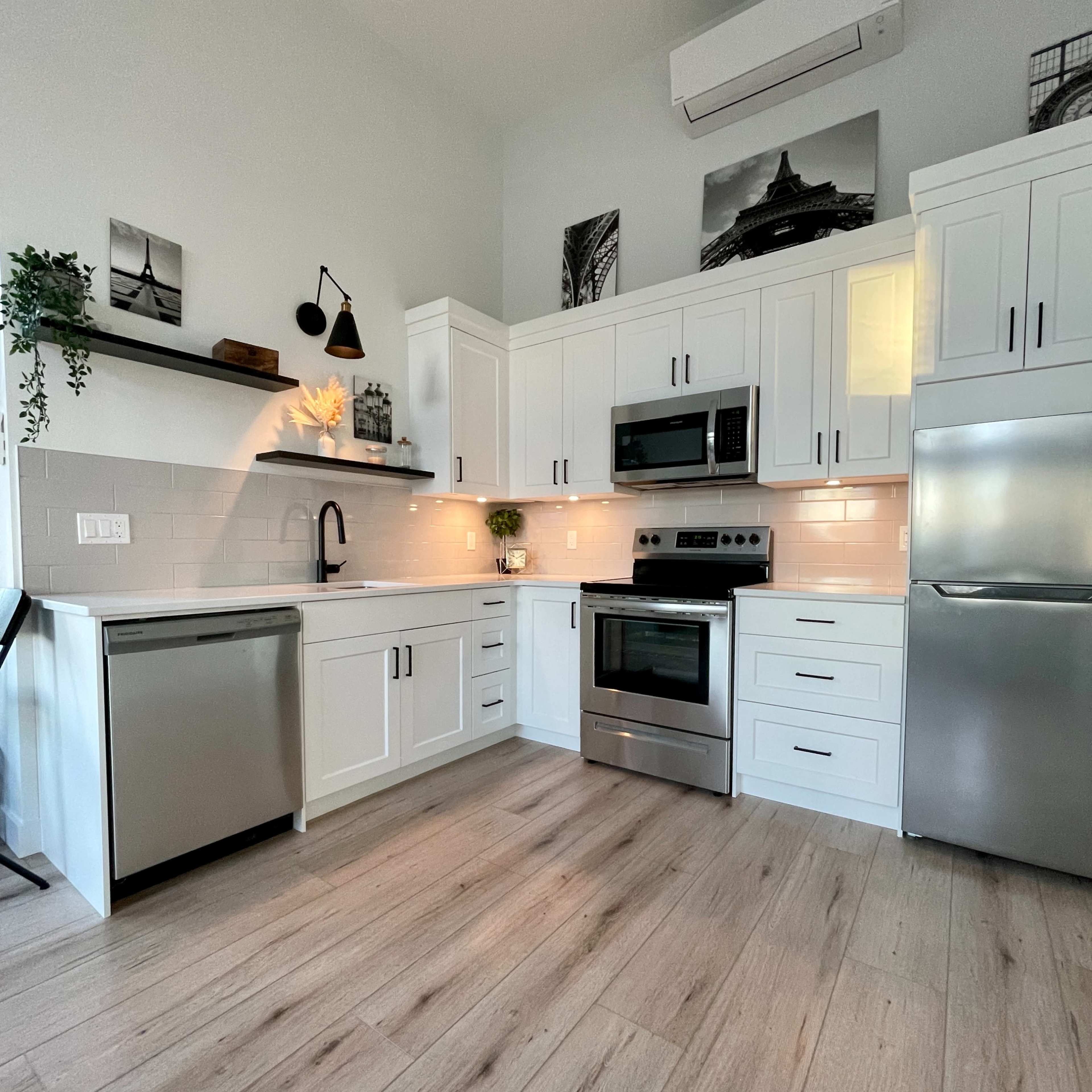 The kitchen features white cabinets, stainless steel appliances, and a light wood floor, with minimalistic decor on the walls.