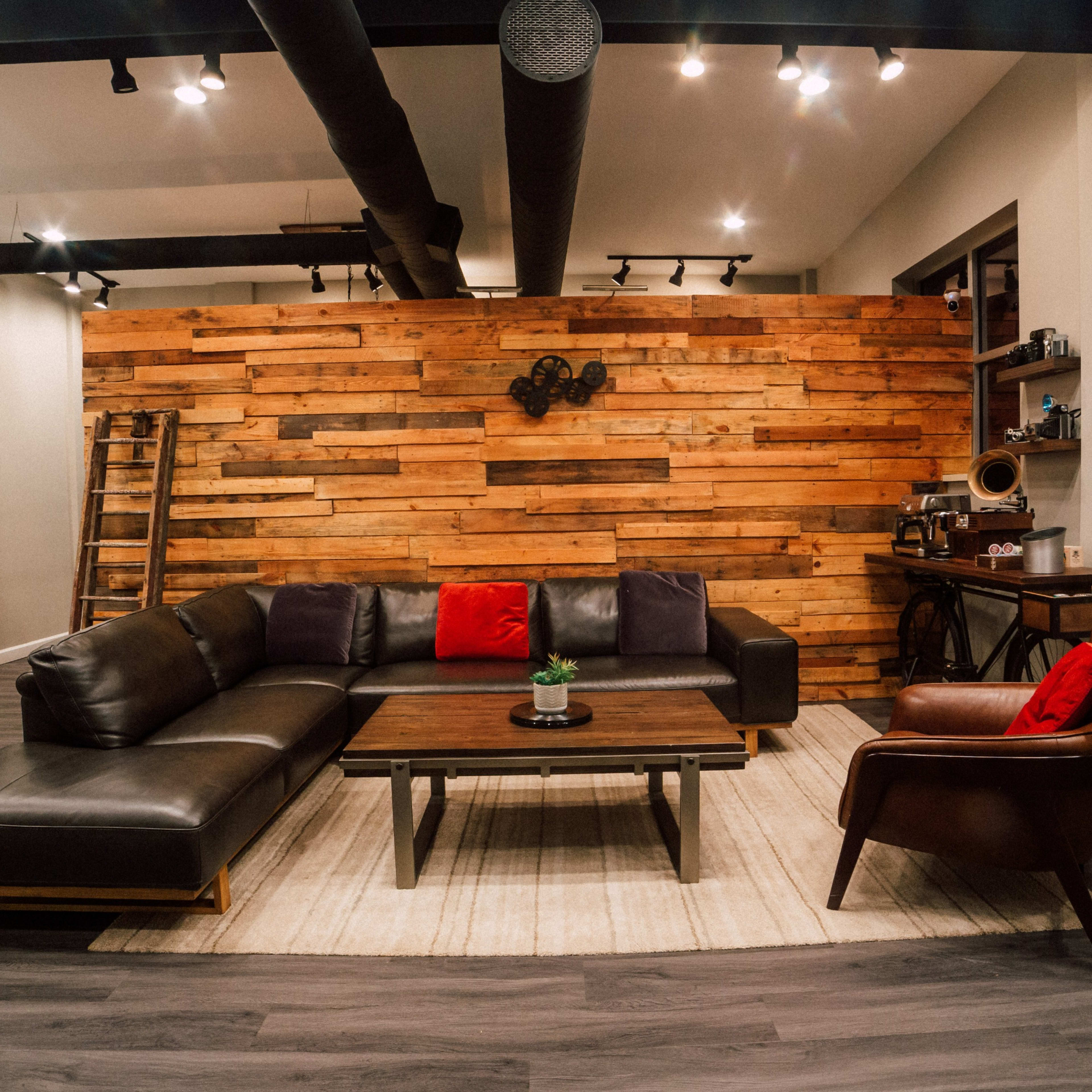 A modern living room features a dark leather sectional sofa with red cushions, a wooden coffee table, and a rustic wood accent wall.