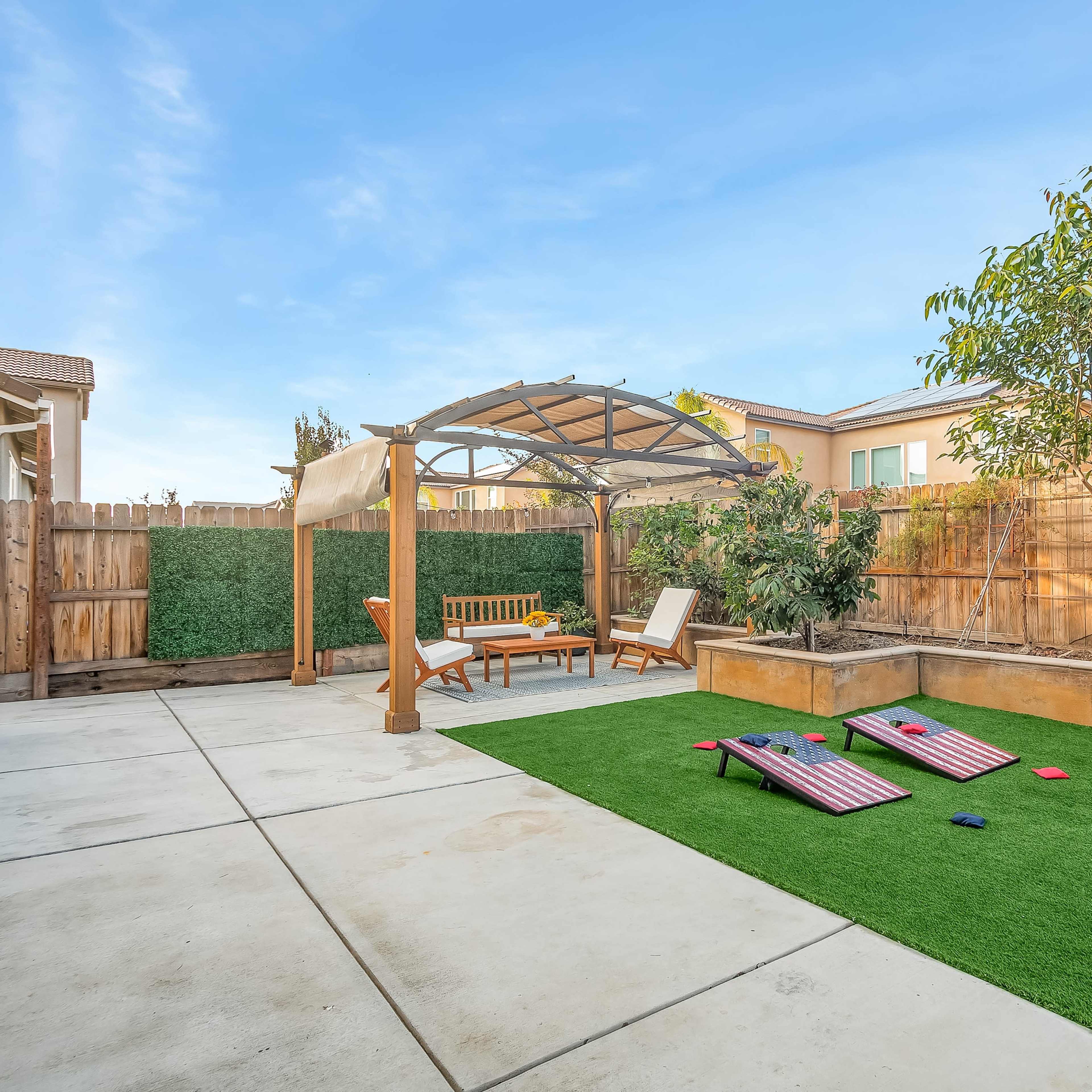 A backyard featuring a pergola, patio furniture, a small lawn area, and cornhole boards on artificial turf.