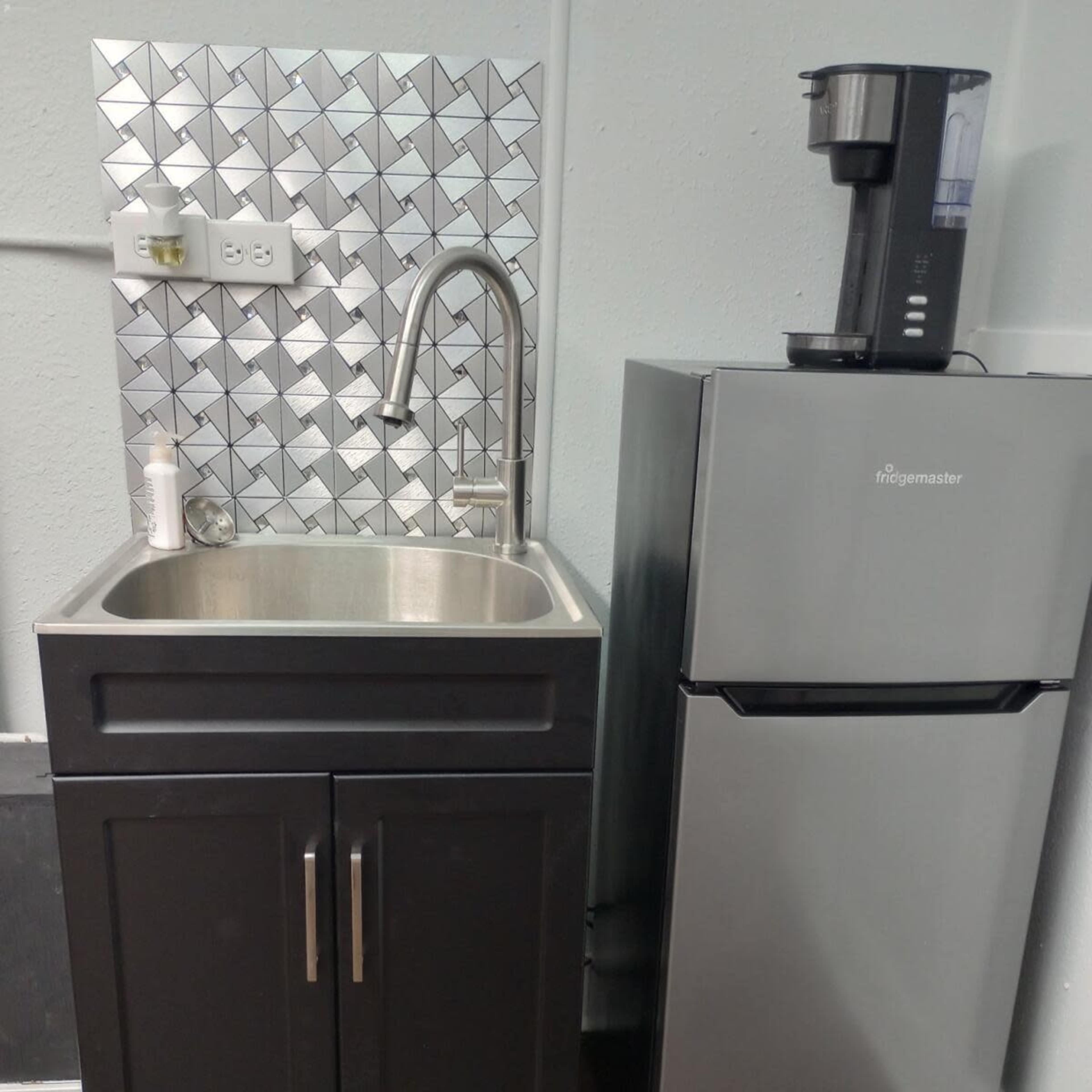 A small kitchen area with a stainless steel sink and black cabinets next to a silver refrigerator and a coffee maker on the counter above.