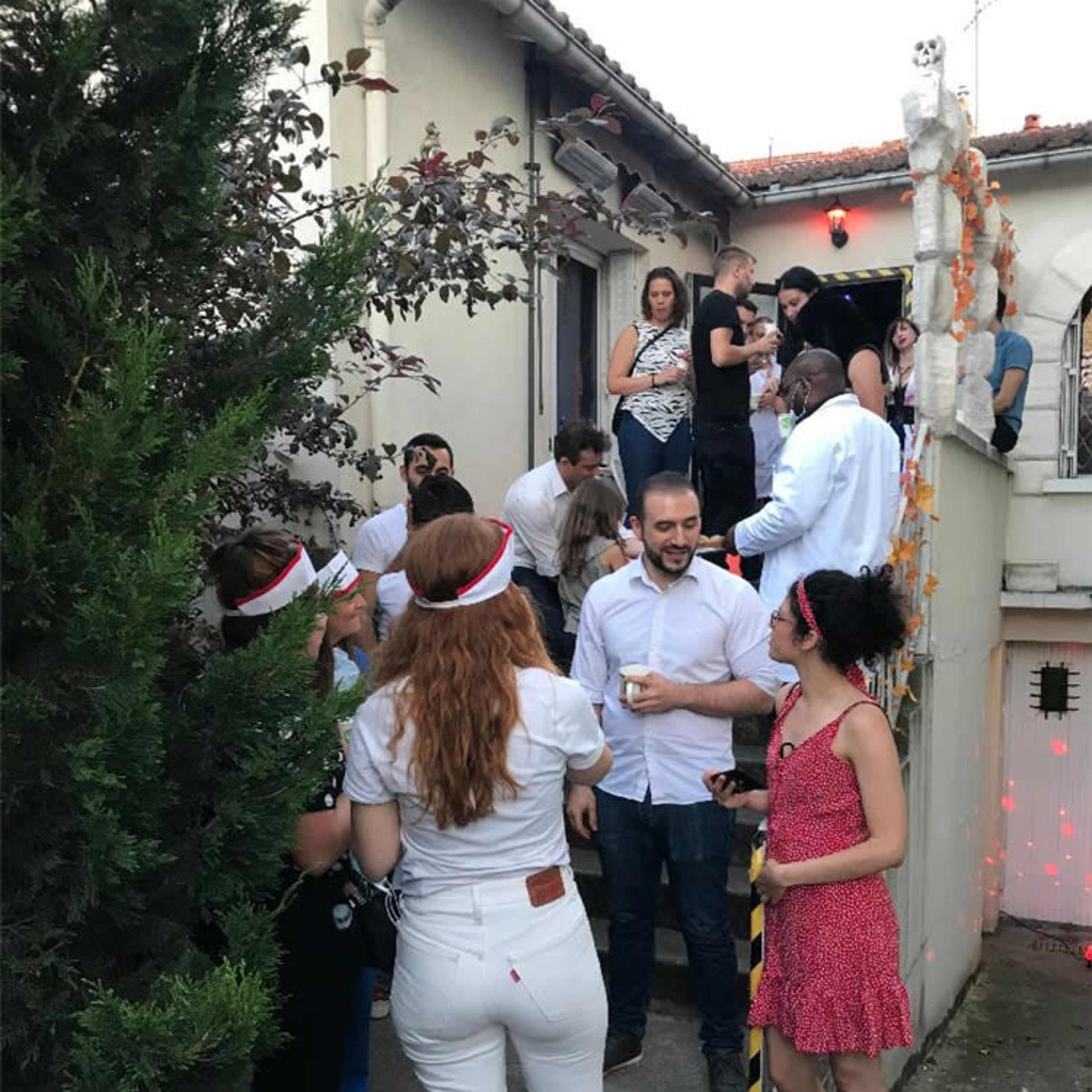 A group of people is gathered outside a house, some on a staircase and others on the lawn, while a few more are engaged in conversation near the entrance.