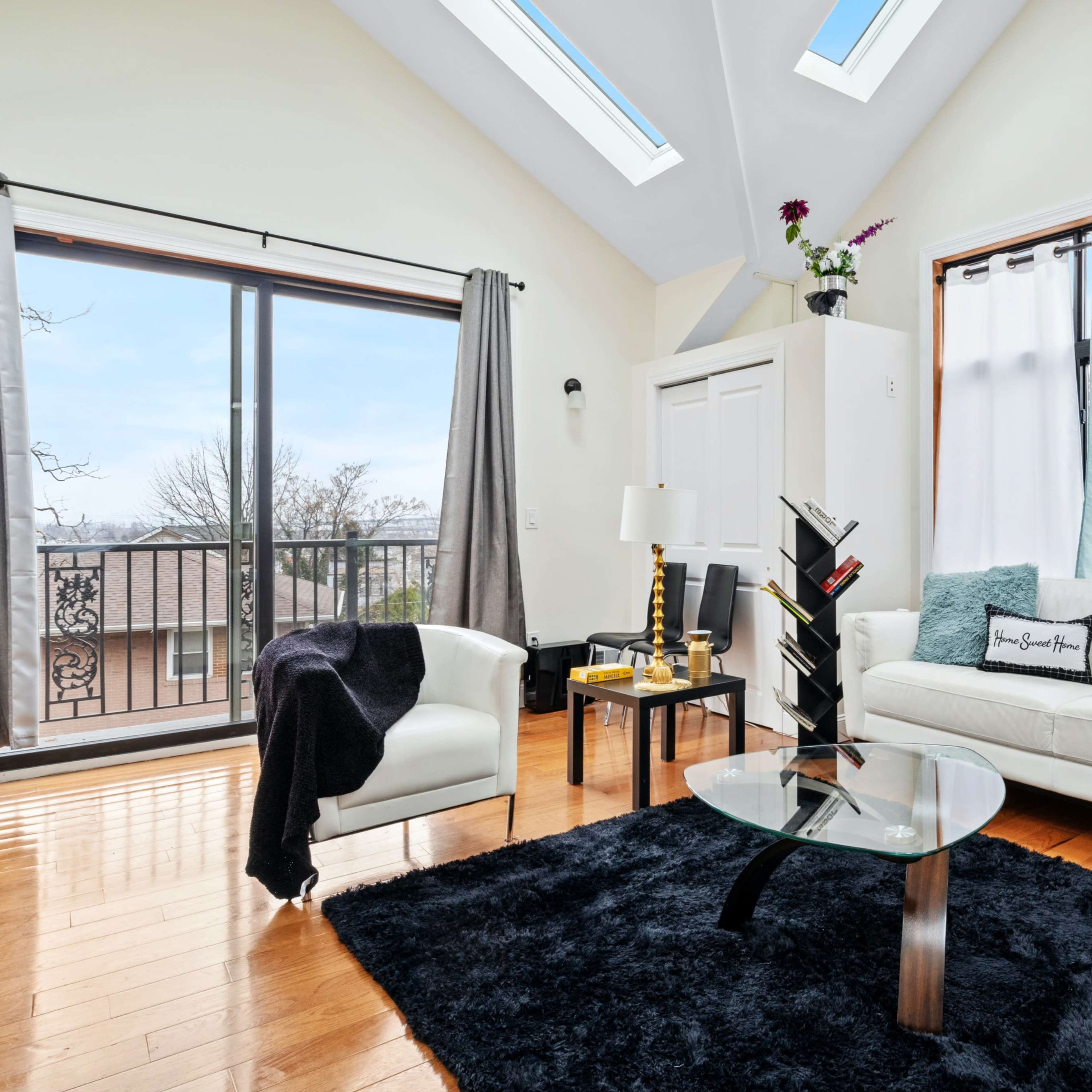 A bright living room features a white sofa, a black rug, large windows with views, and modern decor against a backdrop of wooden floors.