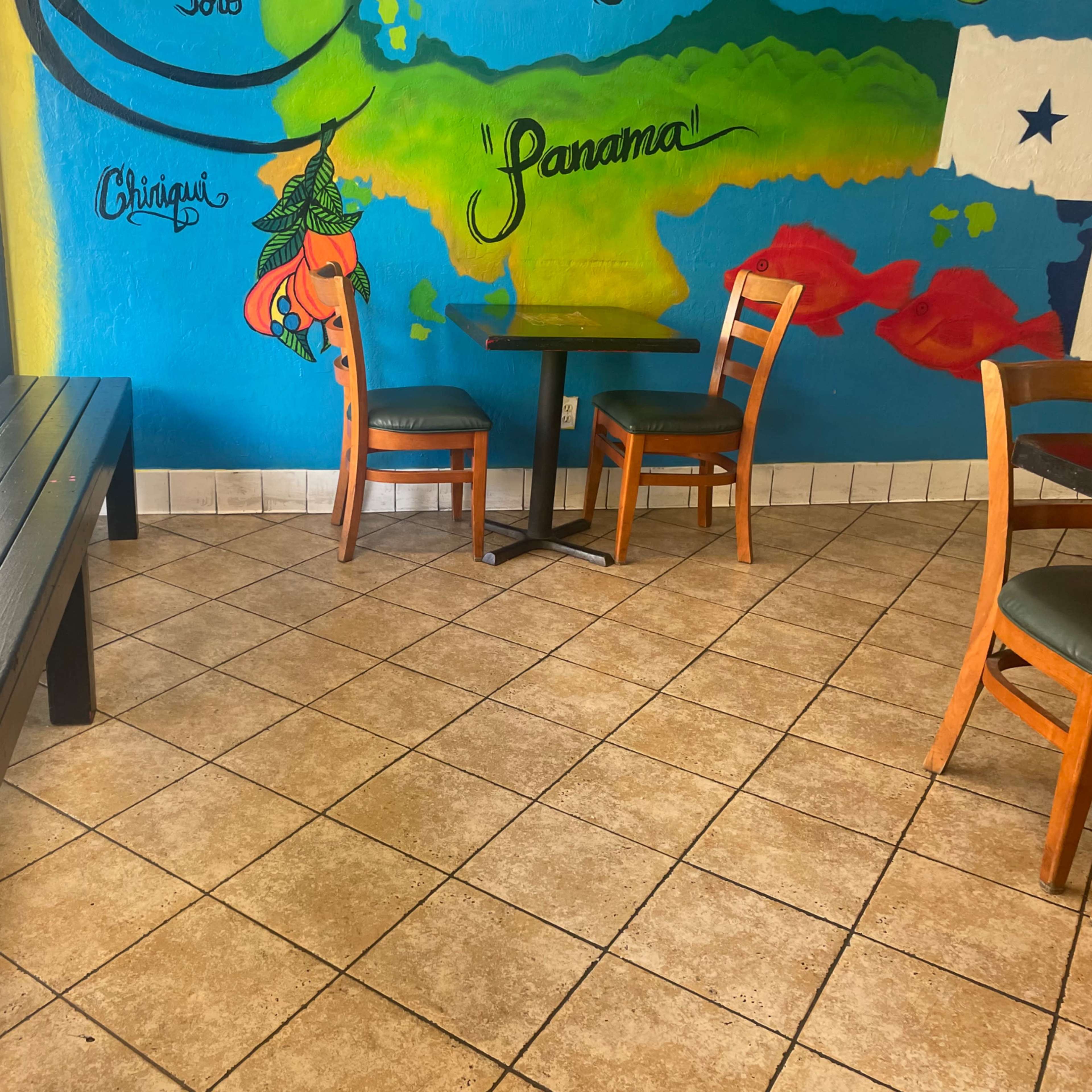 A small dining area with a bench, a table, and three wooden chairs against a colorful mural on the wall.