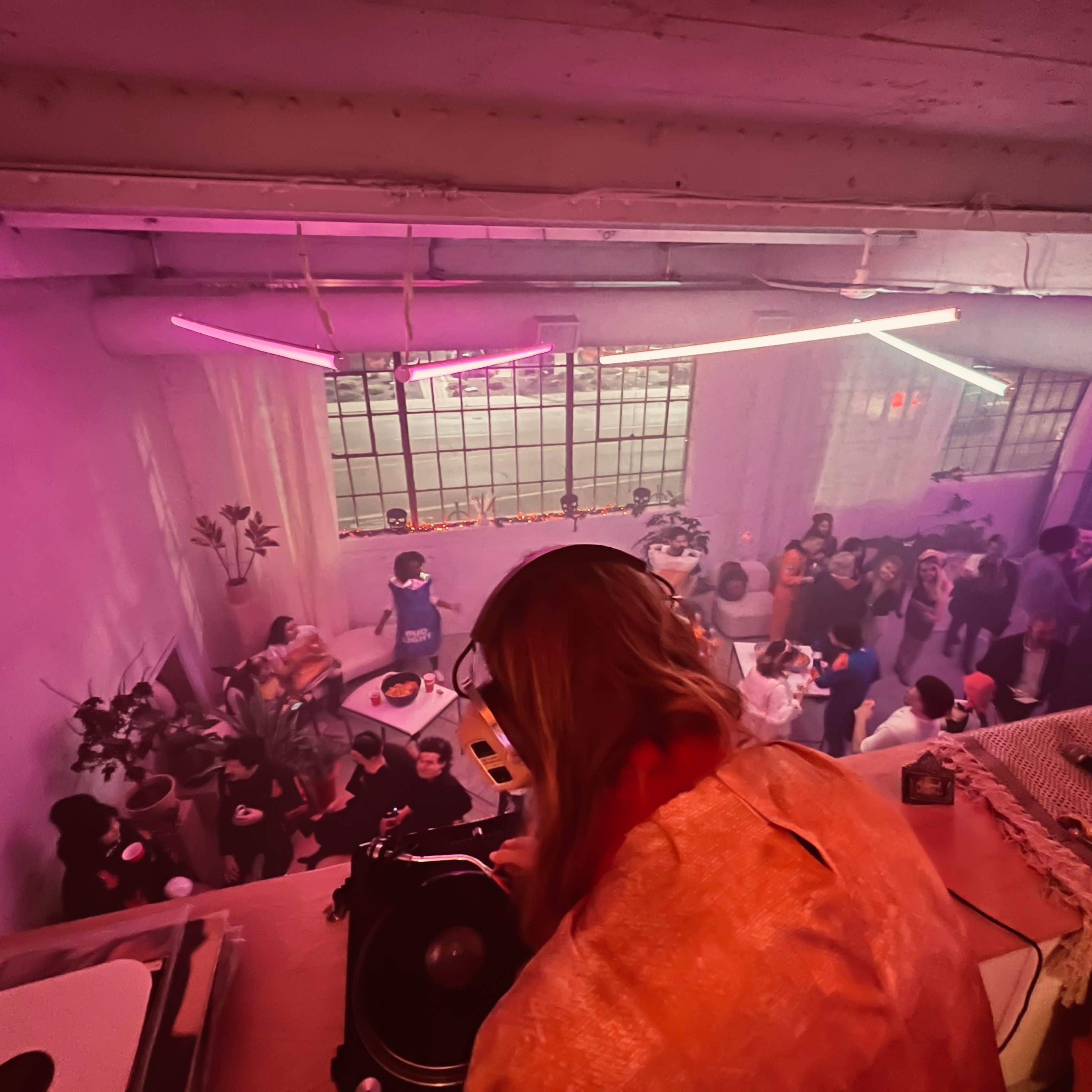 A DJ performs from an elevated position while a crowd gathers below in a dimly lit space decorated with neon lights.