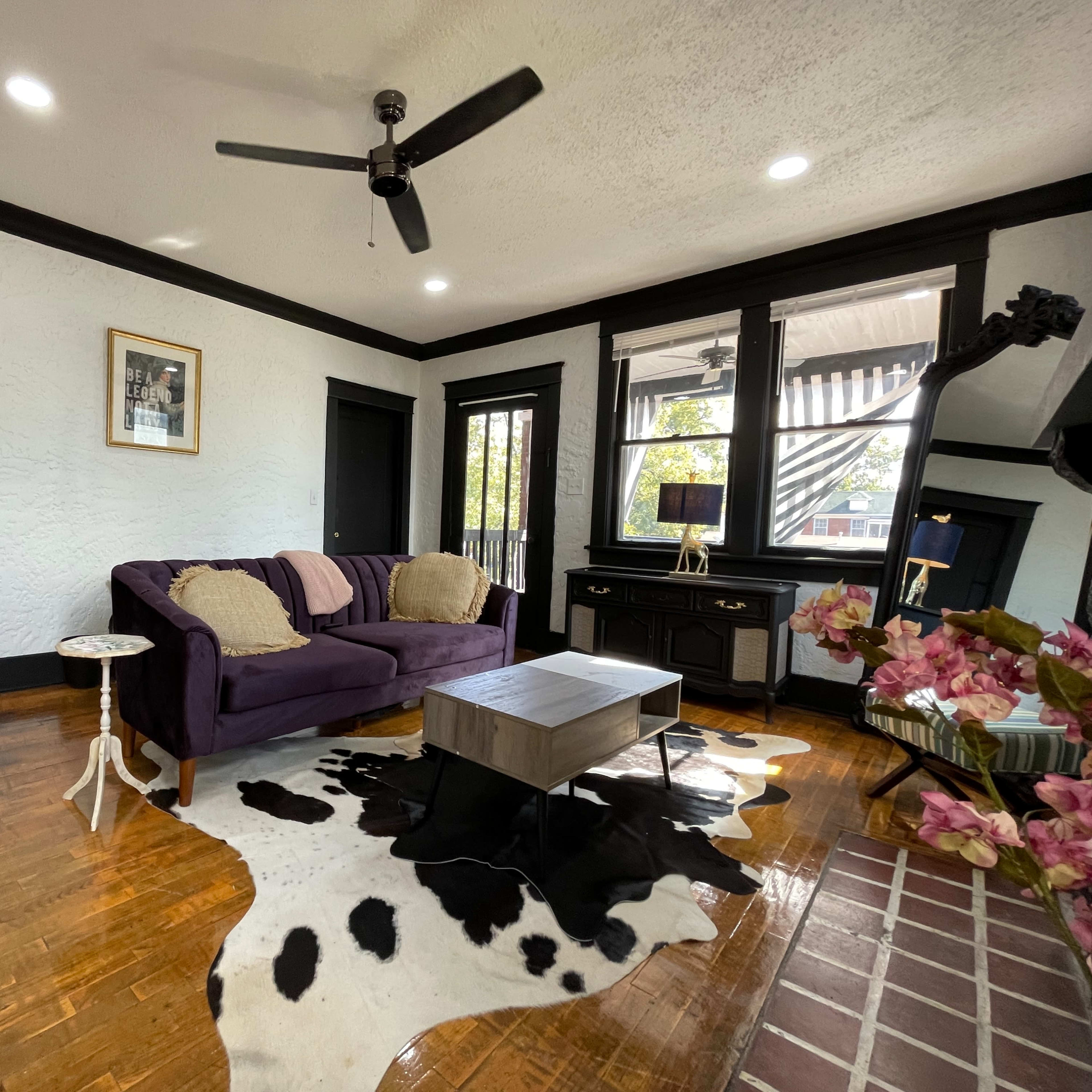 The living room features a purple sofa, a coffee table on a cowhide rug, and decorative elements against a backdrop of light walls and black trim.