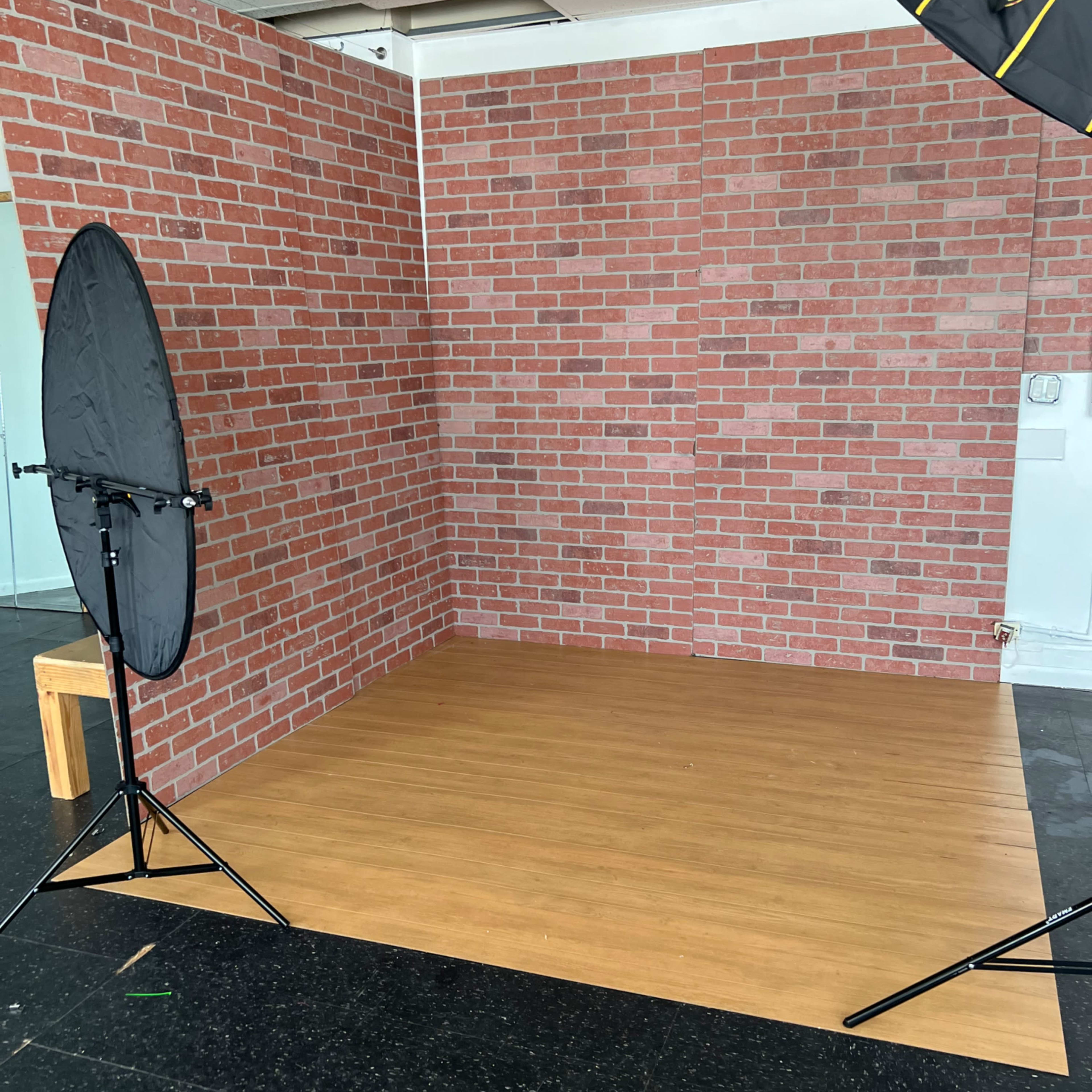 A photography studio with a wooden floor, a faux brick wall backdrop, and two softbox lighting setups.
