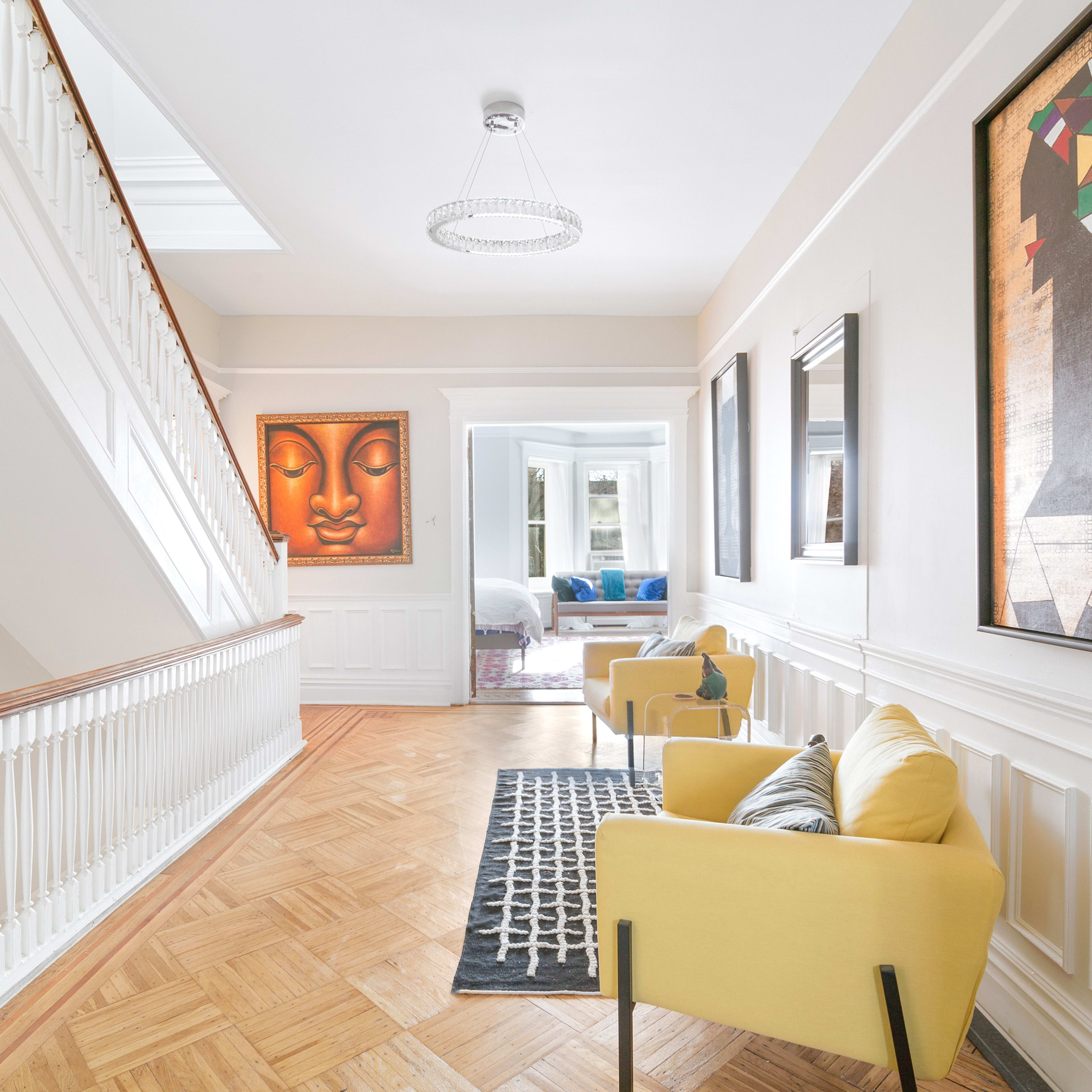 A bright hallway features a staircase, two pieces of framed art on the walls, and two yellow chairs on a patterned rug.